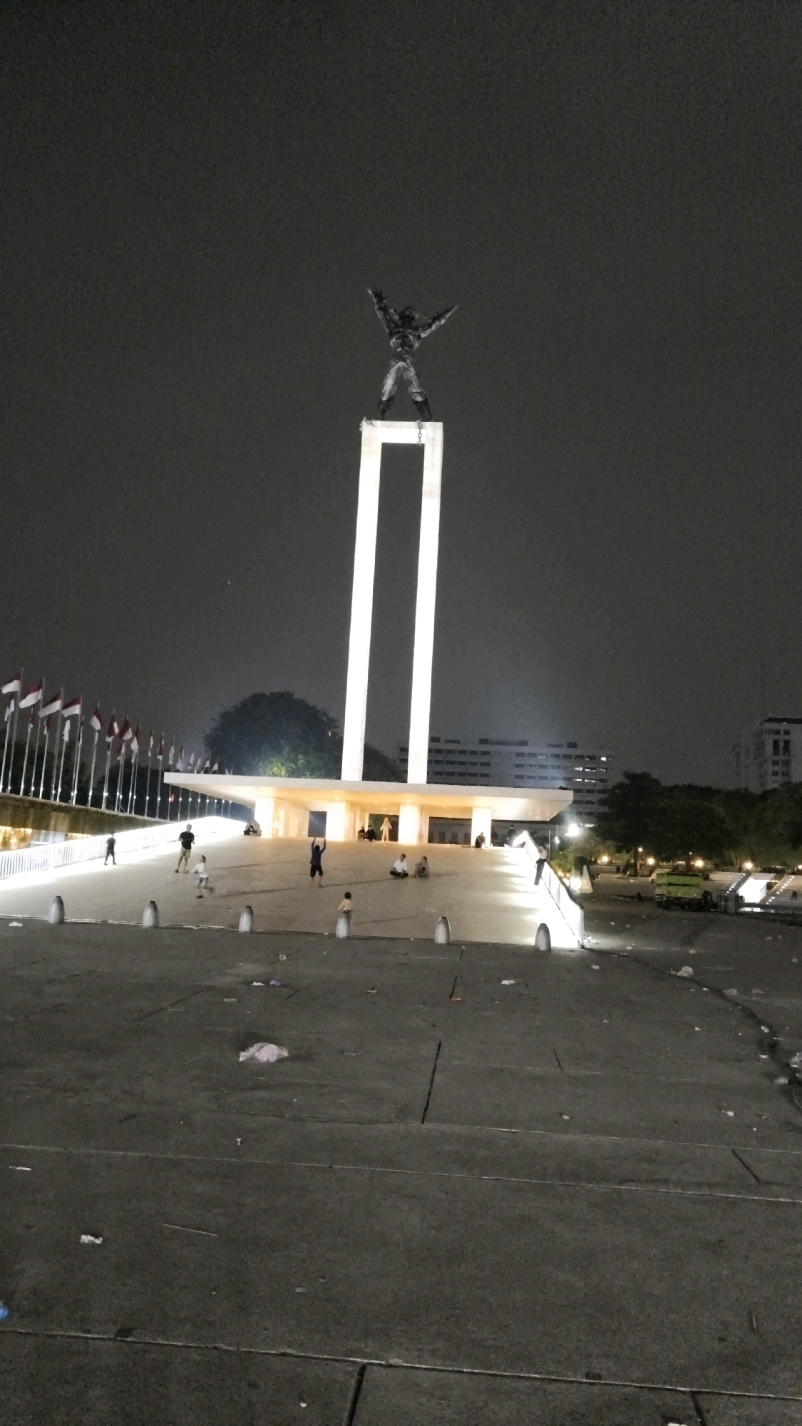 The image depicts the National Monument (Monas) in Central Jakarta, Indonesia, at night. The monument, a tall obelisk topped with a statue of a man with outstretched arms, is illuminated by bright lights. In front of the monument is a large plaza area with a white, tiered structure. Several flags are displayed along the left side of the plaza.
There are a few people visible in the plaza. Some appear to be walking, while others are sitting or standing around the monument. In the foreground, the plaza floor is visible and appears to be littered with trash. A few lampposts are also present, providing additional lighting. In the background, buildings and trees are visible, silhouetted against the night sky. A green truck is parked to the right of the plaza.