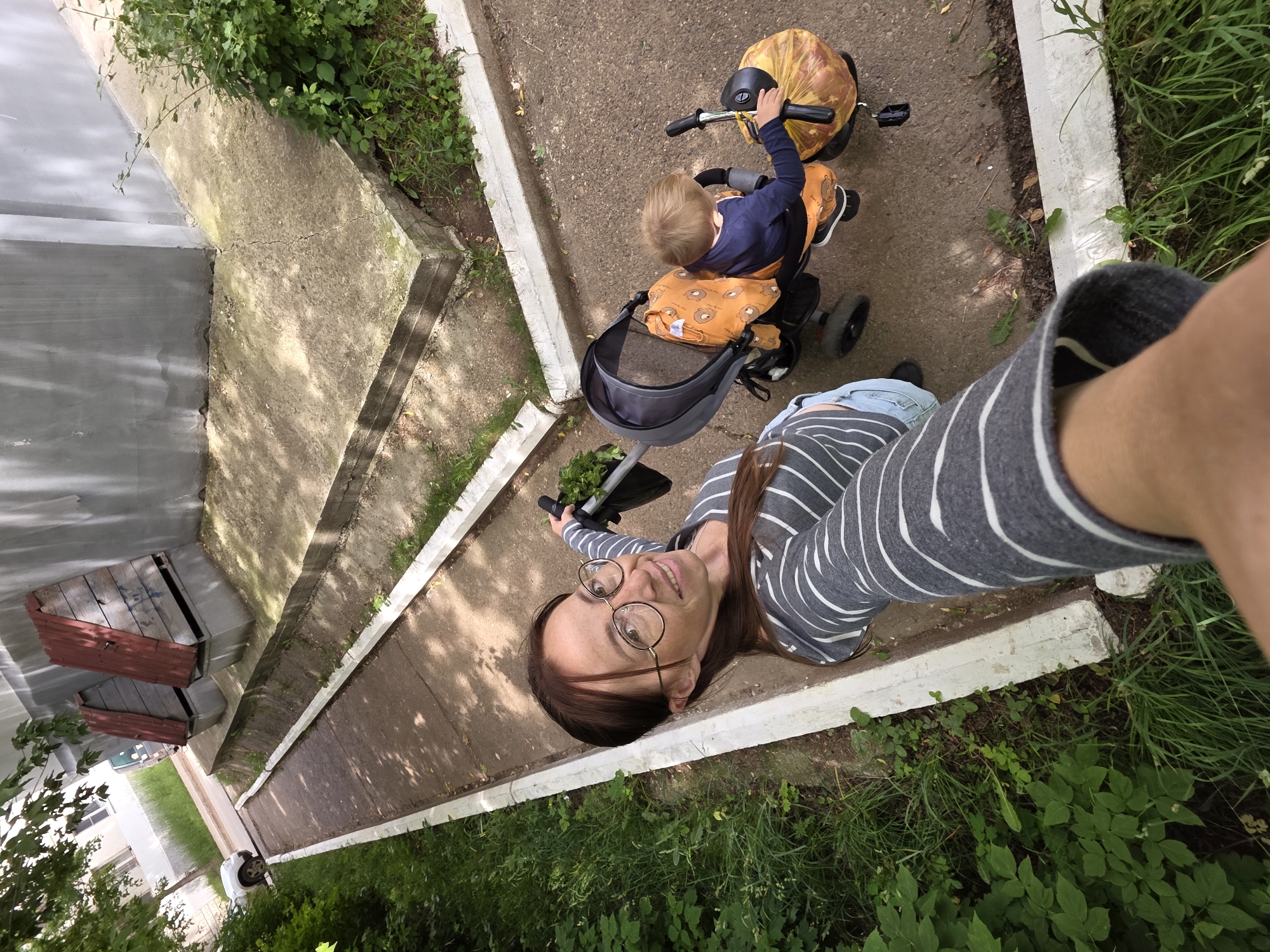 A woman with glasses and a striped shirt takes a selfie while pushing a stroller with a child in it. The child is wearing a dark blue shirt and an orange bib with animal prints. The stroller has a gray canopy and a yellow bag filled with produce. They are on a paved path next to a grassy area with some trees. In the background, there are two wooden structures that appear to be trash bins. The scene is set in a residential area of Chaikovsky, Russia, as indicated by the overall urban environment and building architecture.