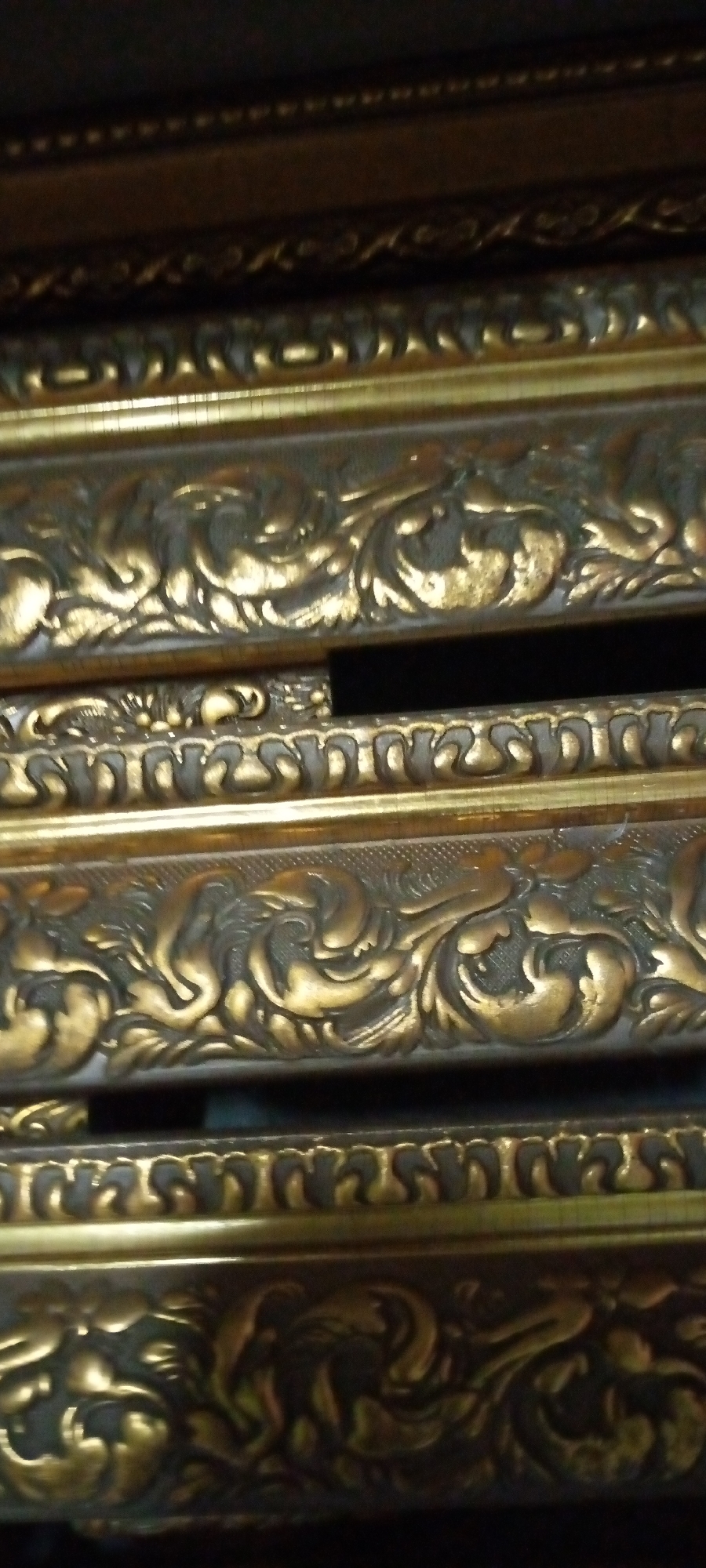 The image shows a close-up, vertically oriented view of several ornate, golden-colored decorative frames stacked one above the other. The frames appear to be made of a material with a metallic finish and feature intricate, carved scrollwork and floral motifs. The lighting highlights the raised details, creating a play of light and shadow that emphasizes the depth and texture of the designs. The background is dark and out of focus, suggesting the frames are in a storage or display setting. There are no people or discernible environmental features of the city of Sumberwono, Indonesia visible in this close-up shot.