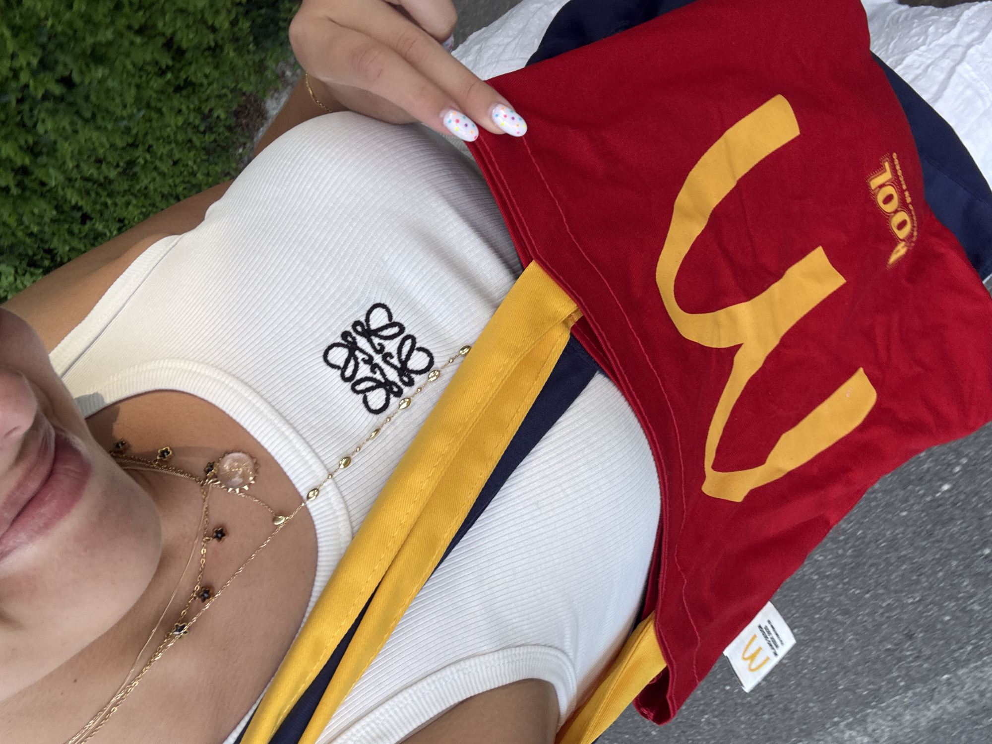 A close-up, first-person perspective captures a person from the chin downward, standing outdoors. The person wears a white ribbed tank top featuring a black embroidered Loewe Anagram logo on the chest. Layered gold necklaces adorn the neck, including one with small black flower charms, another with a clear circular pendant, and a lariat-style chain with small gold beads. A thin gold bracelet is visible on the right wrist. The person's left hand, with white nail polish and colorful polka dot designs, holds a red tote bag with yellow straps. The bag prominently displays the yellow McDonald's "M" logo and, below it, the yellow text "OOL." A white tag attached to the bag shows a smaller McDonald's "M" logo and the text "MILANO DESIGN WEEK 2023," indicating the bag's origin and placing the scene's context in Milan, Italy. A dark blue garment is visible beneath the white tank top. The background is blurred, showing green foliage and a grey paved surface, suggesting an outdoor, daytime setting.