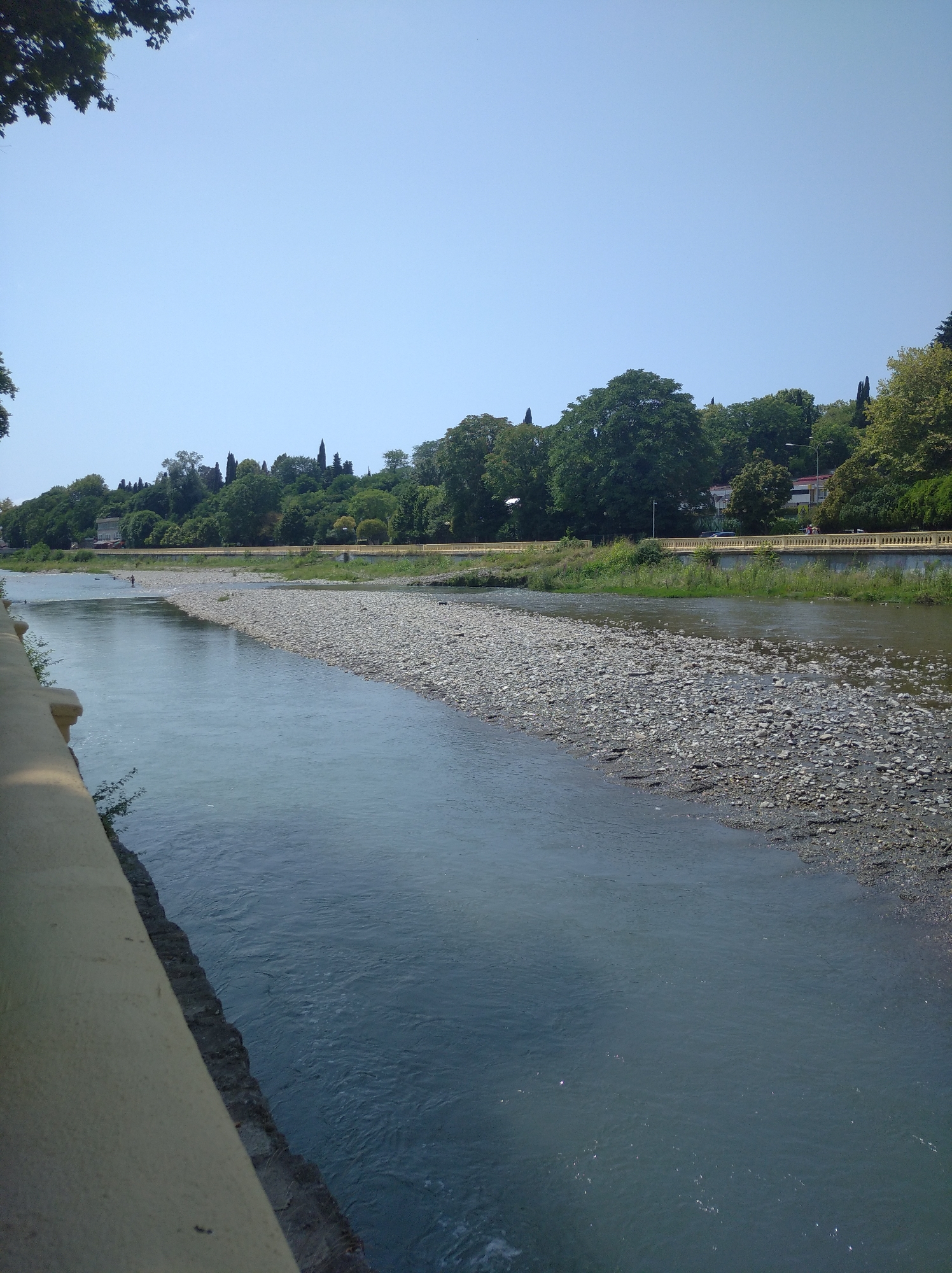A wide river flows through a city. On the left, a yellow-colored retaining wall runs along the riverbank. The river itself is clear and blue, with a visible bed of pebbles and rocks on the right side of the frame. Beyond the river, a grassy embankment is lined with a yellow railing and a pedestrian walkway. Further inland, a dense line of trees forms a green backdrop, with some buildings visible interspersed among the foliage. In the distance, the sky is a clear, light blue. There are a few people visible in the river, as well as a car on the road behind the embankment.