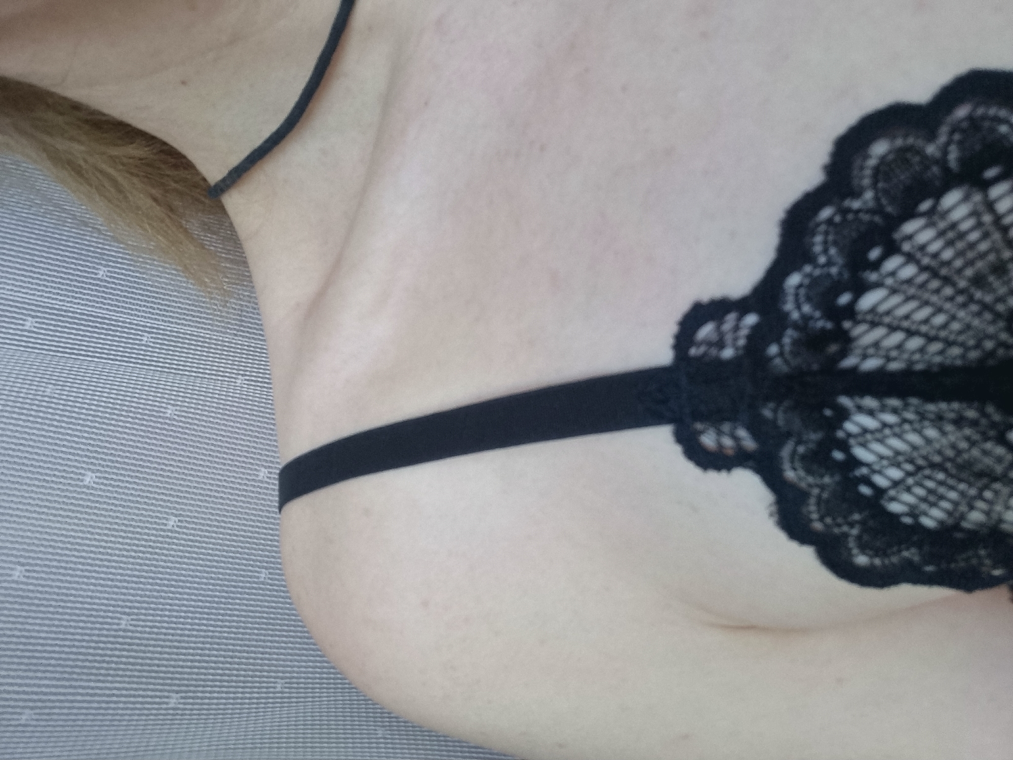 The image presents a close-up, angled view of a person's upper torso, primarily focusing on the chest and shoulder area. The subject's fair skin shows natural texture and a scattering of small freckles or moles. Light-colored hair, possibly blond or light brown, is visible in the upper left corner of the frame, appearing to rest on a surface.
The person is wearing a black lace bralette or bra. The garment features an intricate lace pattern, possibly floral or fan-like, with delicate scalloped edges. A thin black strap of the bralette runs over the shoulder and horizontally across the side of the torso.
To the left and bottom of the frame, a striped fabric surface is visible. This fabric has a fine pattern of parallel horizontal lines, suggesting it could be a pillow, bedding, or upholstery. The overall setting appears to be indoors, likely a private space such as a bedroom or living area.
No specific activity is depicted beyond the person posing for the photograph. The time of day, weather conditions, or specific location within Chaikovsky, Russia, are not discernible from the visual information provided. There is no visible text within the image.