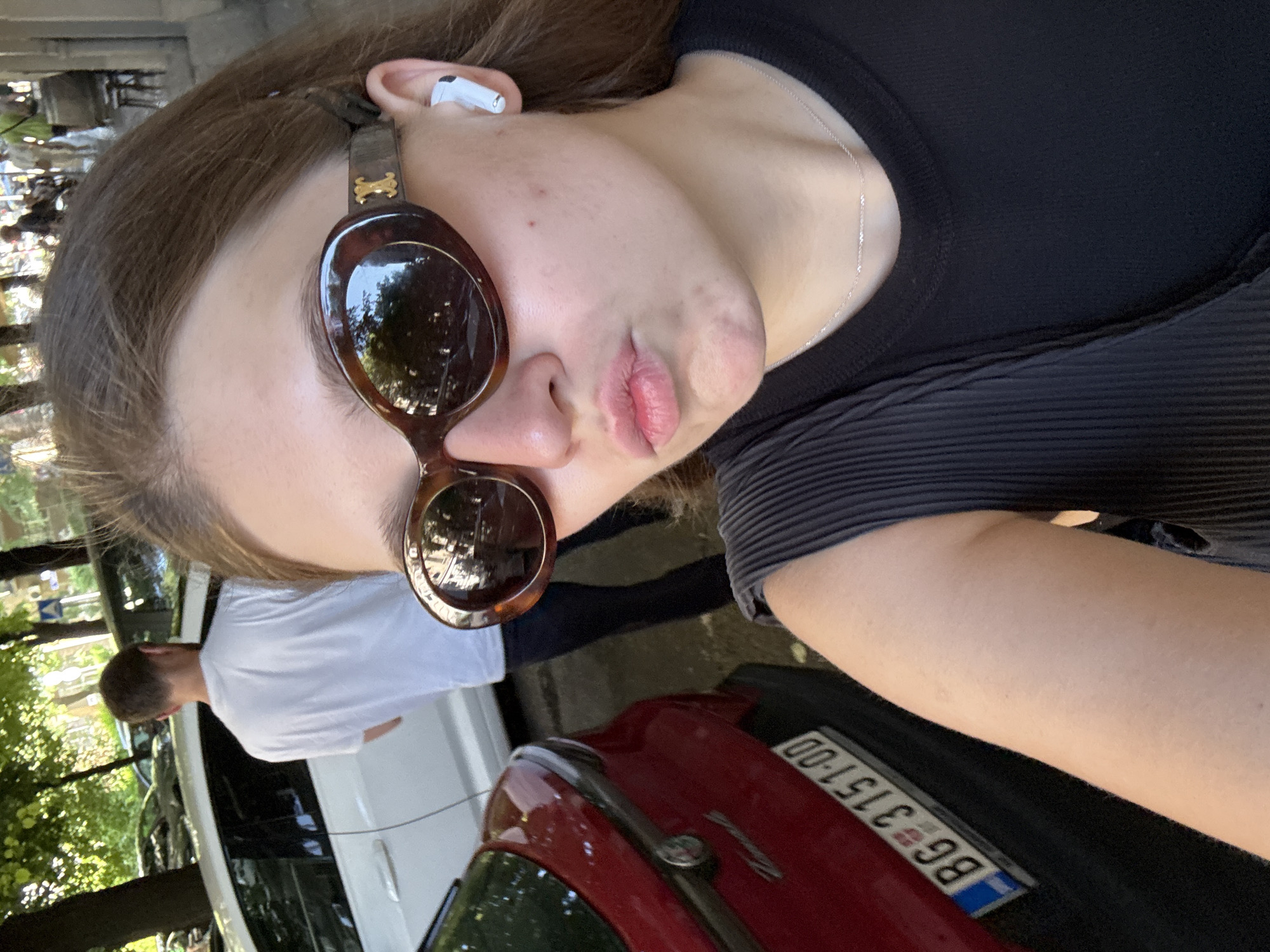 The image shows a selfie of a person with light skin, wearing black tops, sunglasses and an Airpod in their left ear. A silver necklace is visible around their neck. The person is standing next to a red car with a license plate that reads "BG 3151-00." In the background, there is a white car, a man in a white shirt, trees, and buildings. The license plate indicates that the location is in Belgrade, Serbia.