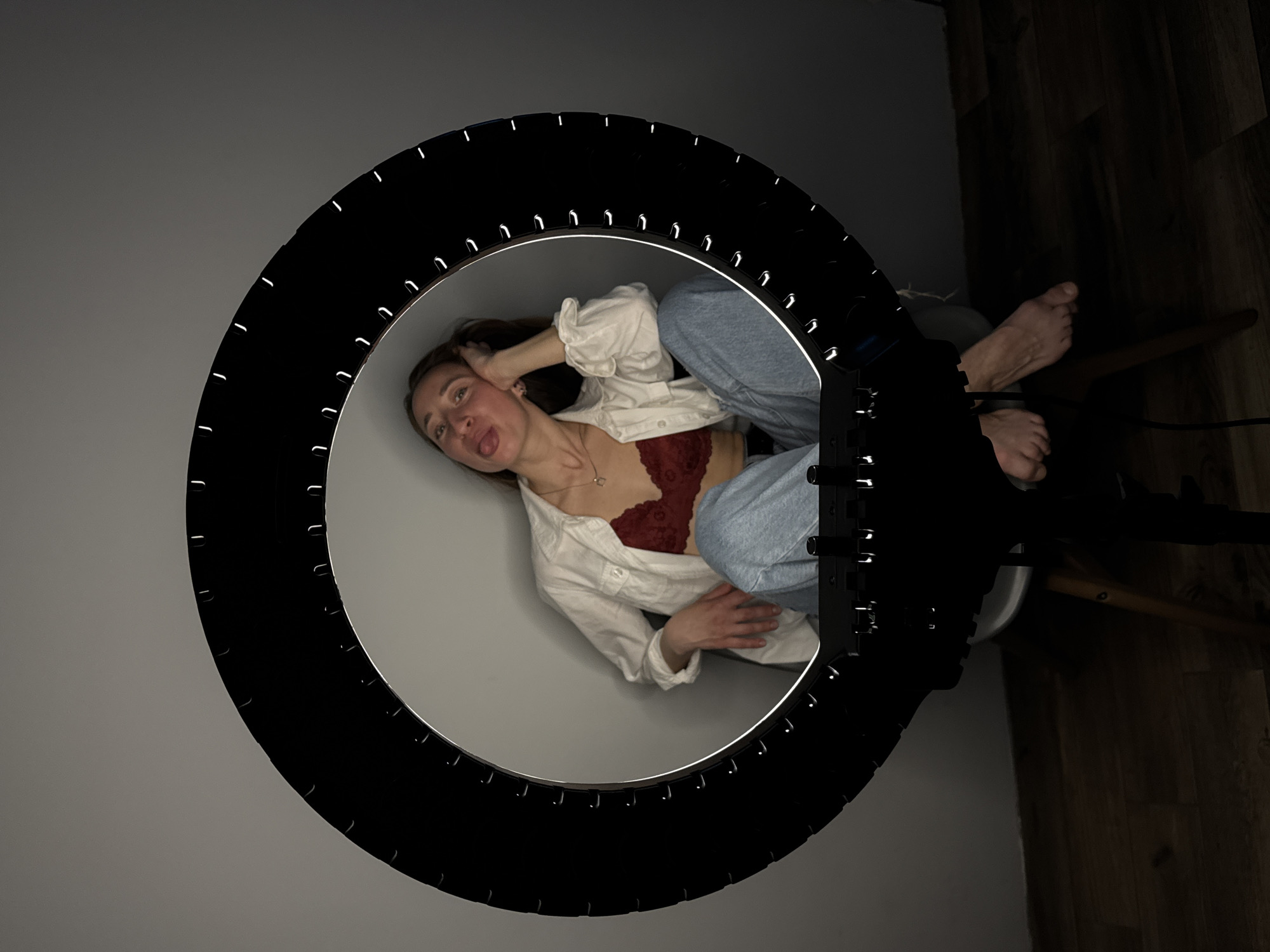A young woman is positioned inside a ring light, sticking out her tongue, wearing a white shirt, a red lacy bra, and blue jeans. She is sitting on a chair. Her feet are visible near the edge of the frame. The floor is dark. The background is a plain, light-colored wall. This image appears to have been taken indoors. There is no information to confirm if this image was taken in Chaikovsky, Russia.