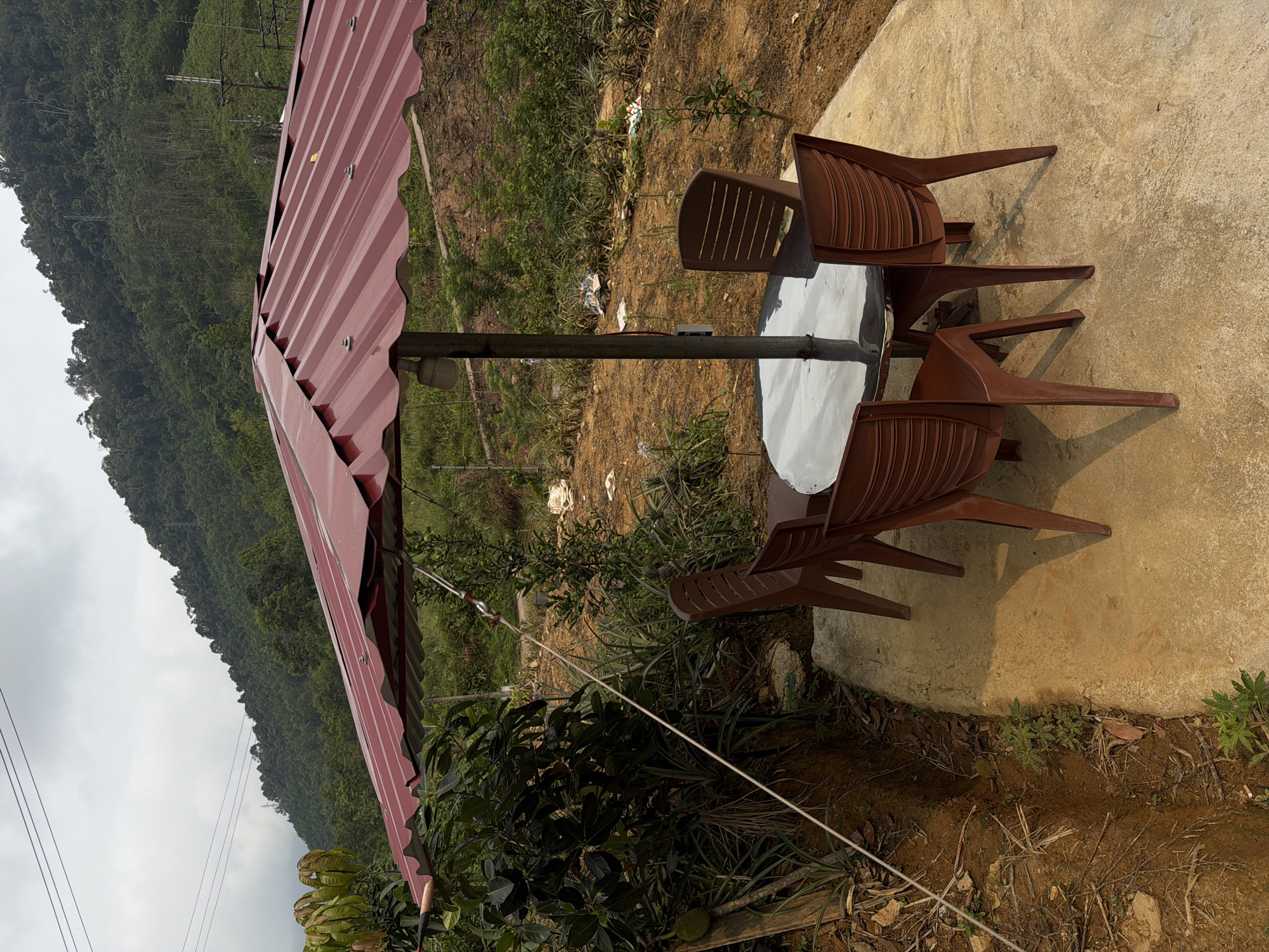 The image shows an outdoor seating area with a metal-roofed structure covering a white, round table surrounded by three brown plastic chairs. The seating area is situated on a concrete slab. Beyond the table and chairs is a view of a hillside covered with vegetation. In the distance, a forested mountain rises. Power lines are visible in the lower left. The vegetation suggests a tropical climate. Without additional information, it is not possible to confirm the location is specifically within the city of Đà Nẵng, Vietnam.