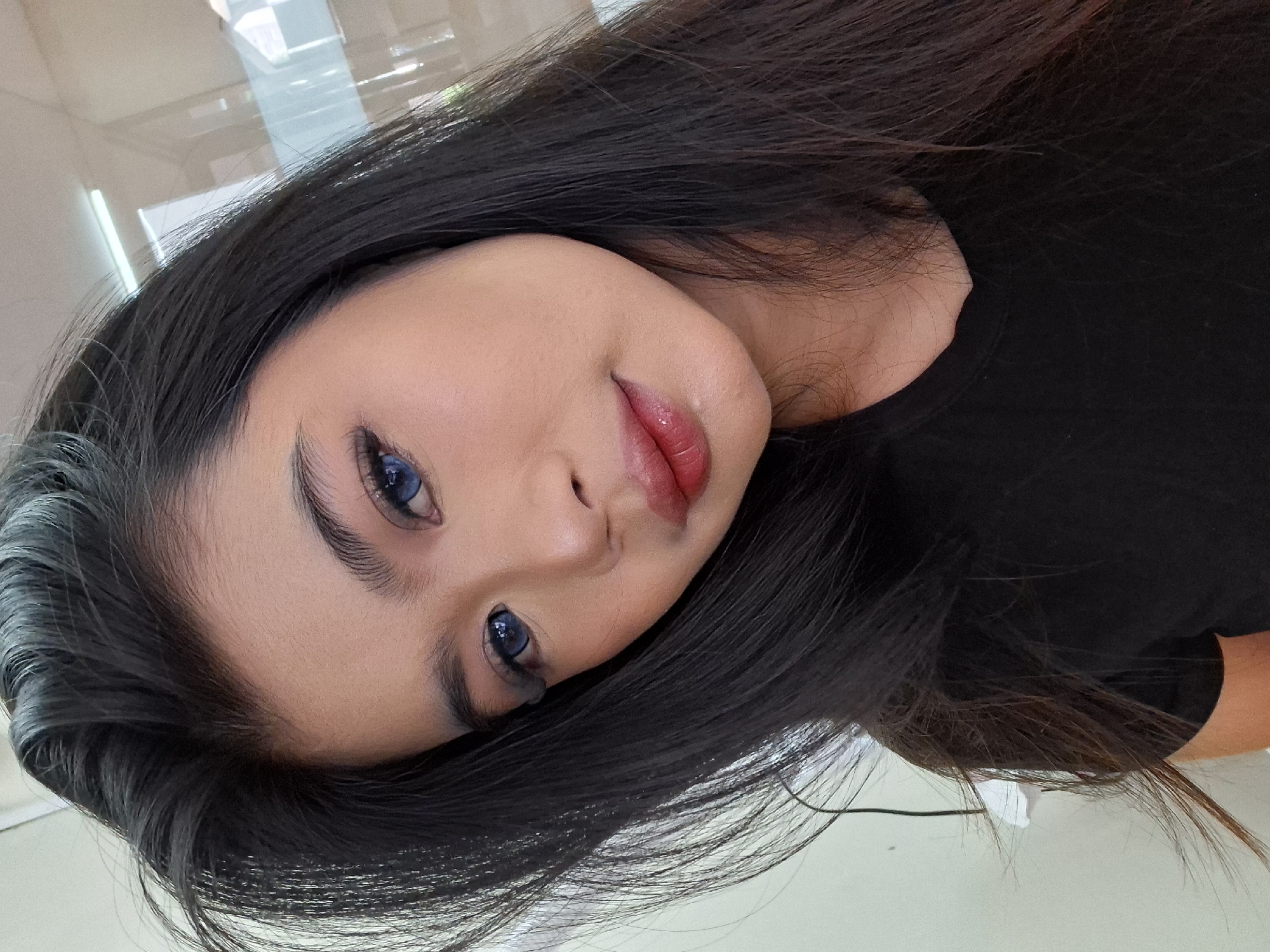 A close-up, head-and-shoulders selfie of a young East Asian woman with long, dark hair, wearing a black t-shirt. She has dark eyebrows, light skin, and appears to be wearing makeup, including blue contact lenses and red lipstick. The background is blurred and suggests an indoor environment with fluorescent lighting and reflective surfaces. The image is taken from a slightly high angle. It is not possible to determine the location in Kuta, Indonesia based on the provided image.