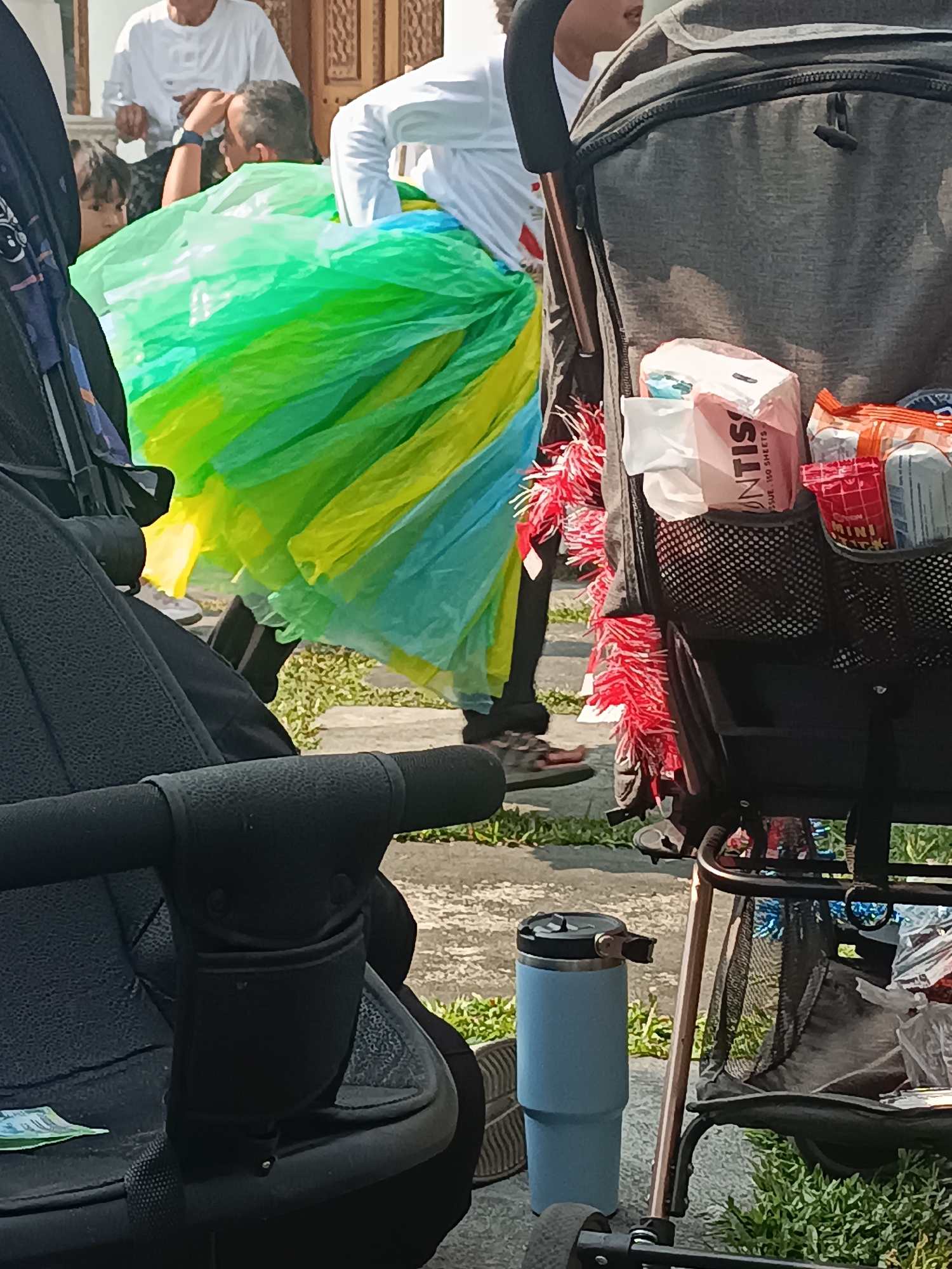 The image depicts a close-up view of two baby strollers, one in the foreground on the left and another on the right. The stroller on the left is black, showing its handle and seat. The stroller on the right is gray, with a mesh storage compartment containing various items, including a pack of tissues, snacks, and other miscellaneous objects. A blue insulated tumbler is placed on the ground next to the stroller on the right. In the background, a person wearing a white long-sleeved shirt and a vibrant green and yellow skirt is visible. Another person in a white long-sleeved shirt is partially visible behind the person in the skirt. The ground appears to be paved with concrete and is partially covered with grass. The scene is set outdoors, likely in a public area. The presence of baby strollers suggests a family outing or event. The location in the city of Sleman, Indonesia, is not directly evident from the visual cues in the image itself, but it is provided as context.