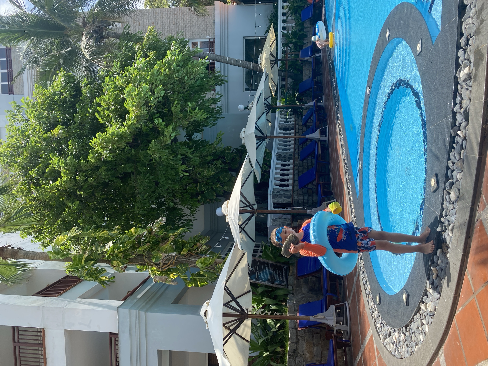 A young boy stands at the edge of a bright blue swimming pool, holding a blue inflatable ring and a grey rock. He is wearing swim trunks and a long-sleeved rash guard. The pool area is surrounded by lounge chairs and umbrellas. In the background, a multi-story building with balconies and lush green trees are visible, characteristic of a tropical resort in Phu Thuy Ward, Vietnam. The sun is shining, creating a bright and clear atmosphere.