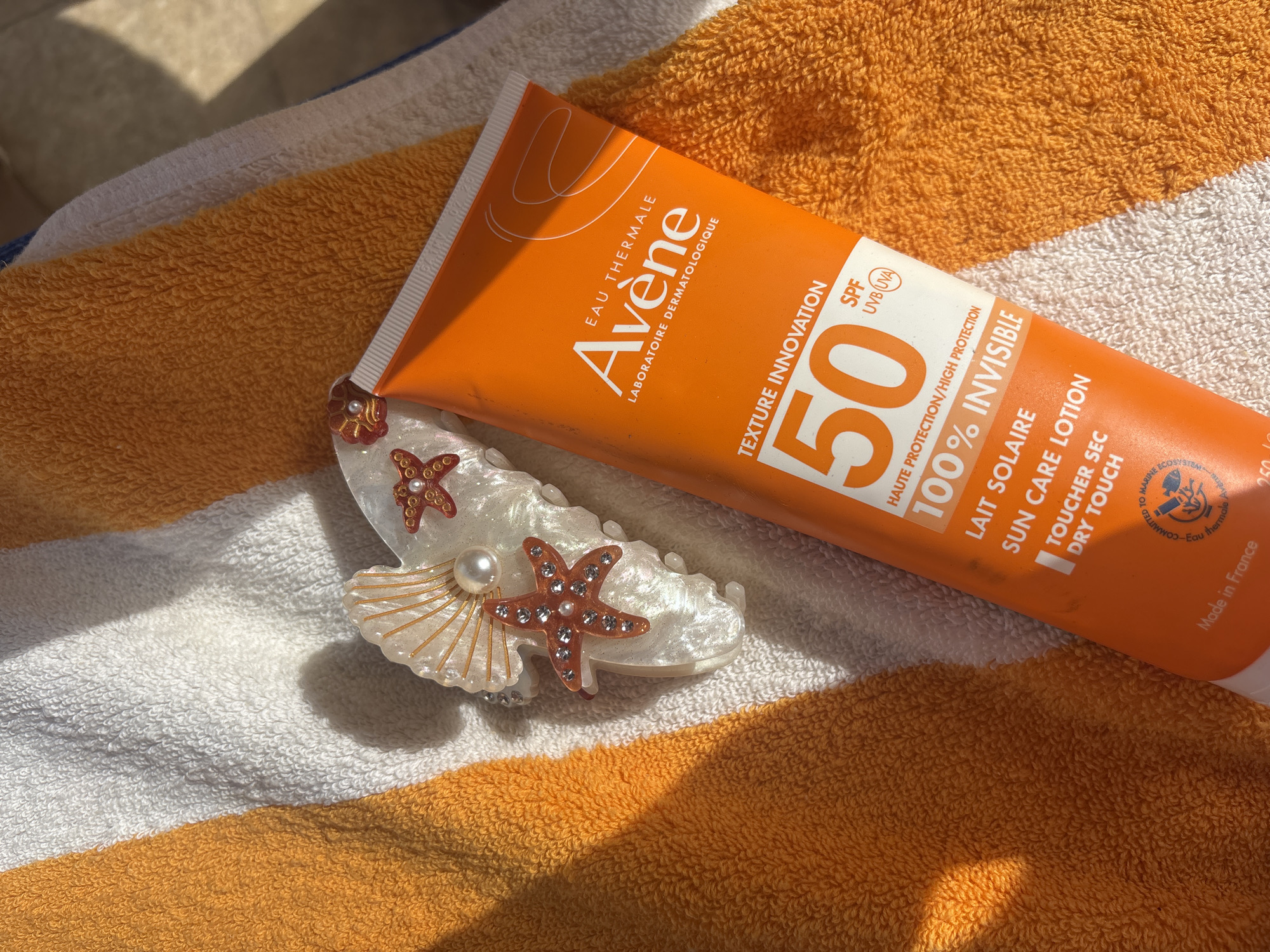 The image shows a tube of Avène SPF 50 sunscreen lying on a beach towel with alternating white and orange stripes. A decorative hair clip, adorned with imitation pearls and starfish-shaped jewels, sits next to the sunscreen tube. The scene is bathed in sunlight, suggesting an outdoor setting, possibly a beach or poolside location. The sunscreen is an orange tube with white lettering, advertising its high protection and dry touch properties. Based on the content, the image was likely taken during a vacation in Sharm El Sheikh, Egypt.