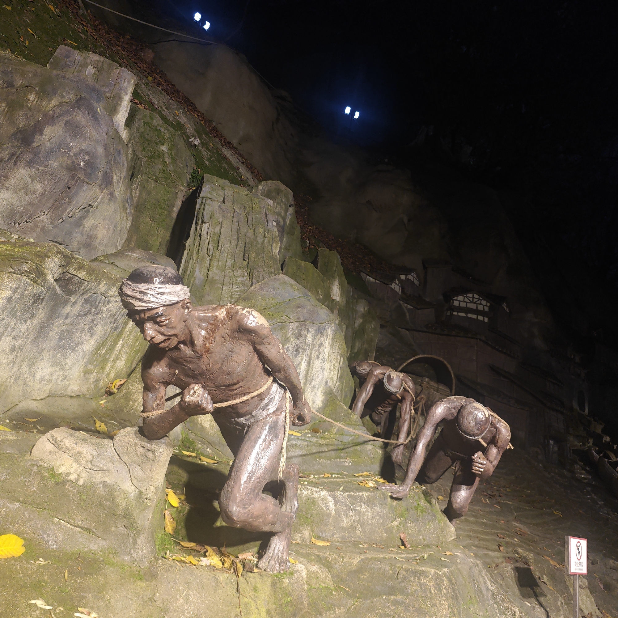 The image depicts a sculpture in Yuzhong District, China, showing three figures pulling a heavy load. The figures are dressed in simple garments and have ropes tied around their waists, which are used to pull the load. The sculpture is set against a backdrop of rock formations. Above the sculpture are bright lights. A sign with Chinese characters is also visible.