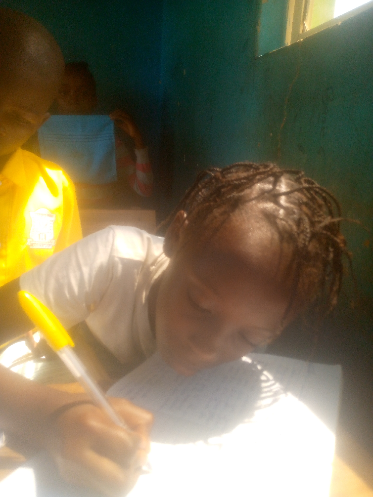 The image shows a young person writing on a piece of paper with a yellow pen. The individual has braided hair and is wearing a white shirt. Another person is visible in the background, wearing a yellow shirt. A third person is also in the background holding up a light blue rectangular object. The scene appears to be indoors, with a green wall and a window visible. The location is Unknown, Unknown.