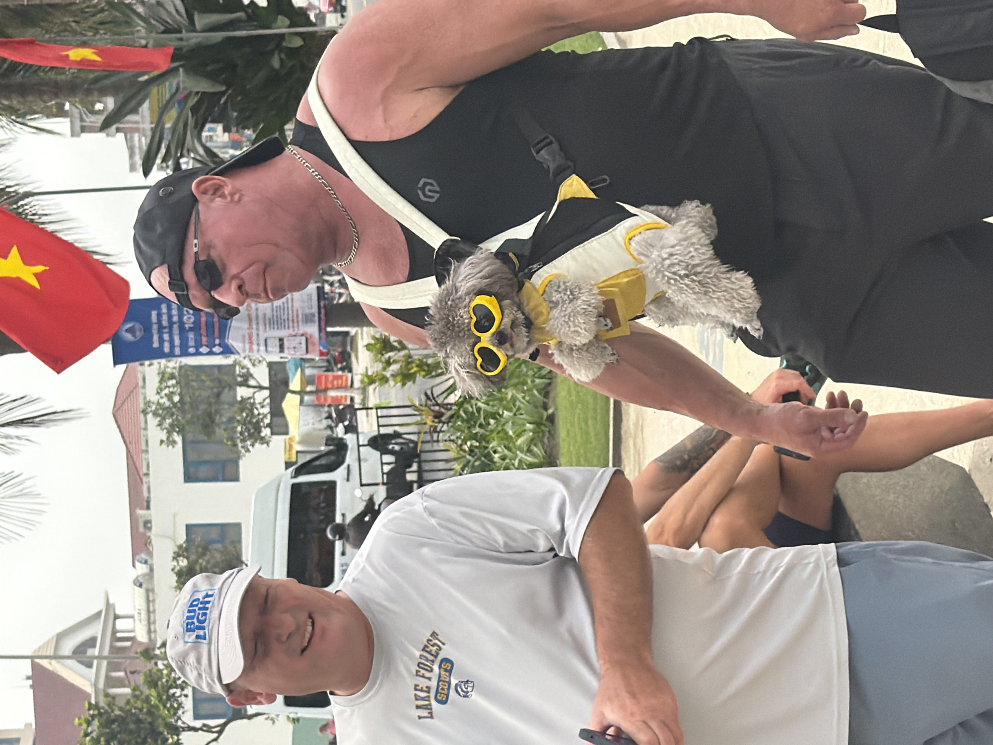 The image, oriented vertically, captures an outdoor scene in Đà Nẵng, Vietnam, on a bright, sunny day. Two men and a small, stylish dog are the primary subjects.
In the upper half, a muscular man with a neutral expression wears a black tank top, black shorts, sunglasses, and a black baseball cap worn backward, along with a silver chain necklace. He is carrying a fluffy gray dog in a black and white sling-style carrier across his chest. The dog is a notable detail, dressed in a yellow and white outfit and distinctive yellow heart-shaped sunglasses.
In the lower half, another man, smiling slightly, wears a light gray t-shirt with "LAKE FOREST SCOUTS" and a logo, and a light gray baseball cap featuring "BUD LIGHT." He holds a smartphone in his right hand. The legs and arms of a third person, wearing dark blue shorts and appearing to be relaxing or seated, are partially visible in the lower right corner.
The background includes light-colored buildings with red roofs and blue window frames, lush green foliage including palm trees, and a white van. Two vibrant red Vietnamese flags with yellow stars are prominently displayed on the left side of the image, clearly indicating the location. The overall atmosphere is relaxed and leisurely.