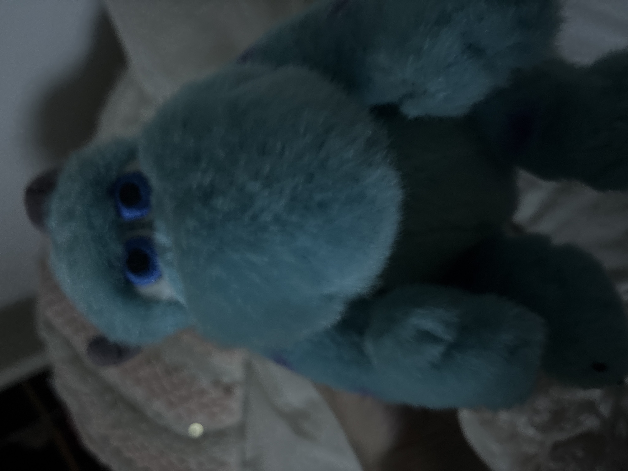 A plush blue stuffed animal with blue eyes is lying on a textured surface. The scene appears to be indoors.