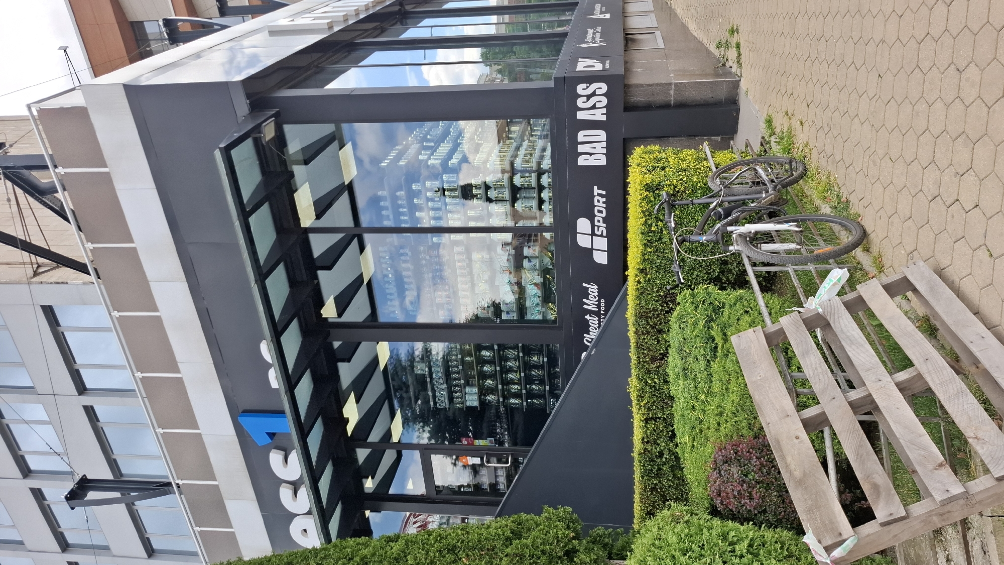 The image shows the exterior of a modern building with large glass windows, reflecting the sky and surrounding trees. The building has signage that reads "ACC", "SPORT", "BAD ASS", and "Cheat Meal - Healthy Food". Inside the store, shelves filled with bottles and products are visible through the windows.
Outside the building, a bicycle is leaning against a metal rack. Next to the rack, a wooden pallet lies on the ground. A neatly trimmed hedge runs along the front of the building, separating it from a paved walkway. The paving stones have a hexagonal pattern.
The lighting suggests it is daytime, with bright sunlight casting shadows, and the sky is partly cloudy, indicating fair weather. The scene appears to be in an urban or commercial area. There are no people visible in the image. The overall impression is of a clean and well-maintained commercial establishment.