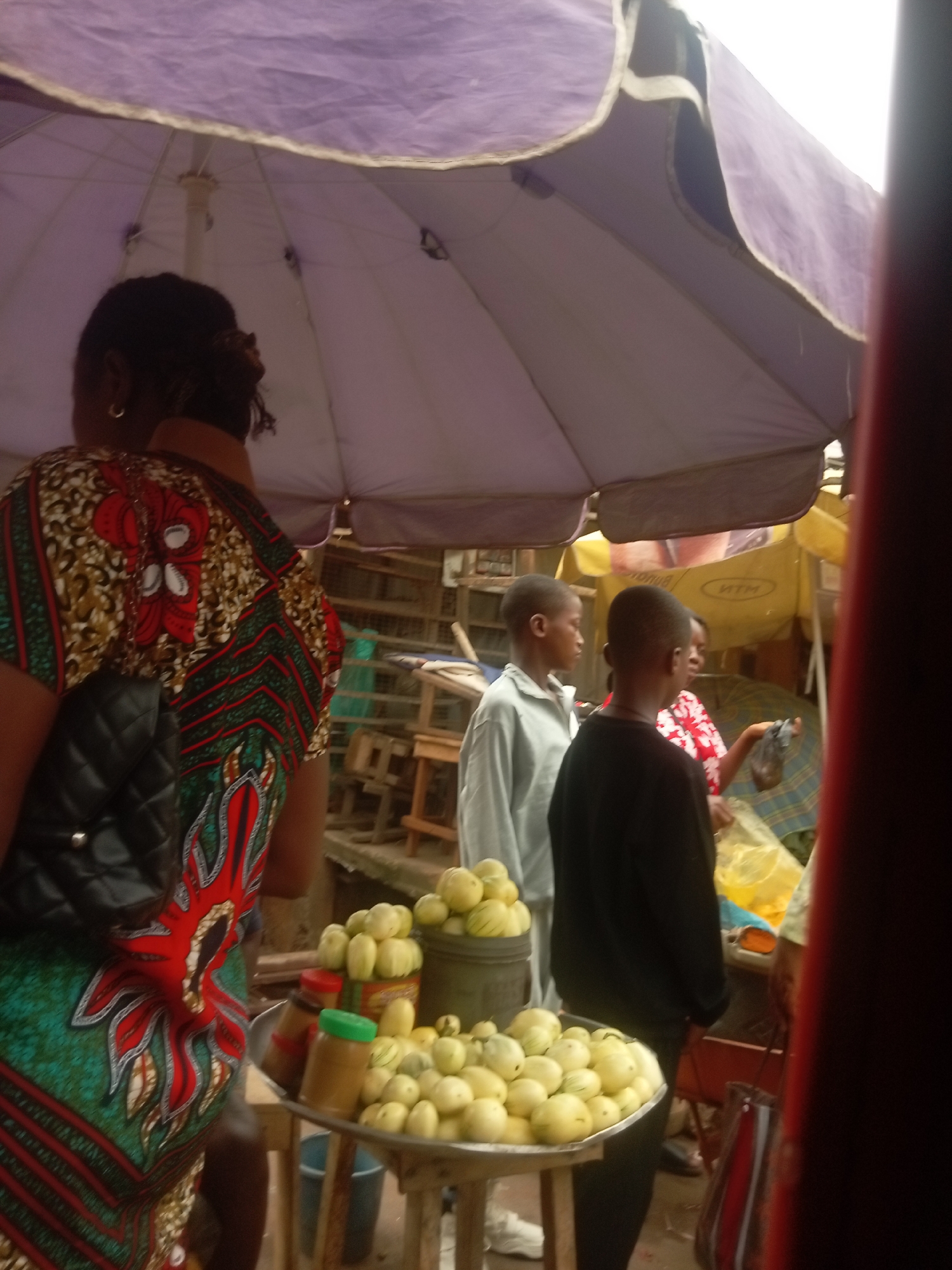 The image depicts a market scene in Nkpor, Nigeria. Under the shade of a large purple umbrella, a woman in a patterned dress and black purse stands near a table laden with yellow produce, possibly melons or gourds, and jars with colorful lids. Two young boys are present, one in a gray shirt and the other in a black shirt. Another woman in a floral print shirt is partially visible, interacting with someone near a yellow MTN-branded umbrella. Various wooden stools and other market infrastructure can be seen in the background.