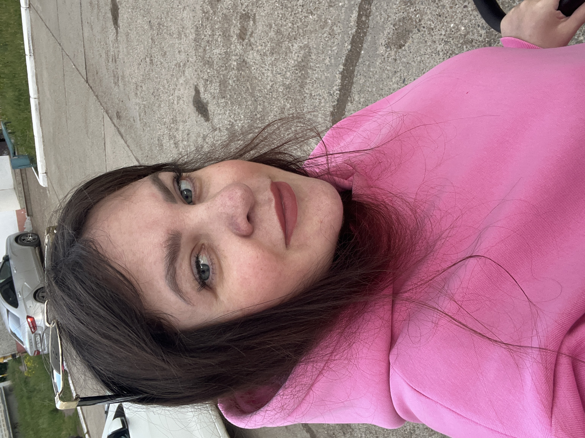 A close-up, portrait-style image features a young woman looking directly at the camera in an outdoor setting in Noviy, Russia. She has dark brown hair that falls around her face and shoulders, and light-colored eyes, possibly blue or grey. Her lips are adorned with a mauvy-pink lipstick. She is wearing a bright pink hooded sweatshirt, which prominently fills the lower and right portions of the frame. A pair of sunglasses with a gold and black frame is resting on her head or hair.
The woman has a calm, neutral to slightly smiling expression. The background reveals a grey concrete or asphalt surface with various textures and cracks, suggesting a paved area like a driveway or sidewalk. Patches of green grass are visible along what appears to be a curb or edge of the paved ground. Parts of several vehicles, including a silver car and a white vehicle, are subtly visible in the background, indicating a residential or urban environment. The lighting suggests it is daytime, with soft, diffused natural light. The overall activity appears to be a casual selfie or portrait taken outdoors. No legible text is visible in the image.