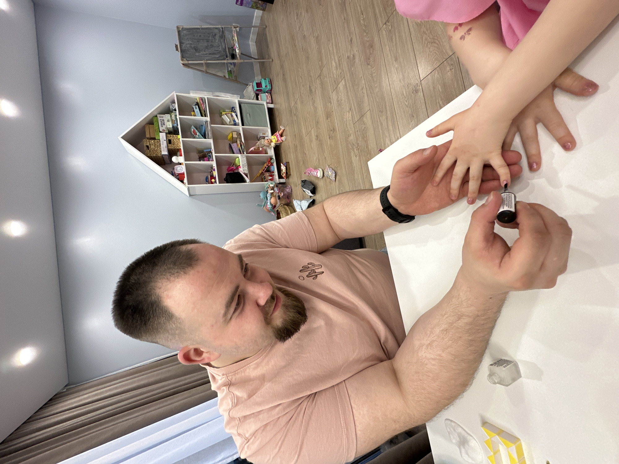 In a brightly lit room, likely a child's play area or bedroom in Chaikovsky, Russia, a man is seen carefully applying nail polish to a child's fingernails. The man, with a short, neat haircut and a beard, is wearing a light pink t-shirt and a dark smartwatch on his left wrist. He is focused on the task, holding a small bottle of shimmery pink nail polish and a brush, meticulously painting the child's nails.
The child's hands are small, with several fingernails already adorned with the pink polish. The child is wearing a pink garment, and a temporary tattoo depicting small butterflies is visible on their left arm. Their hands rest on a clean, white table surface where the nail polishing activity is taking place.
The background of the room features light blue walls. A distinctive white, house-shaped shelving unit filled with various items like books, baskets, and toys is mounted on the wall. Further back, a chalkboard on a stand is partially visible. The floor is light-colored wooden planks, and several toys, including a doll and a pair of pink shoes, are scattered across it. The ceiling is white with bright, recessed lighting fixtures, and brown curtains can be seen along one side of the room. The scene depicts a tender and playful interaction. The label on the nail polish bottle is not clearly legible.
