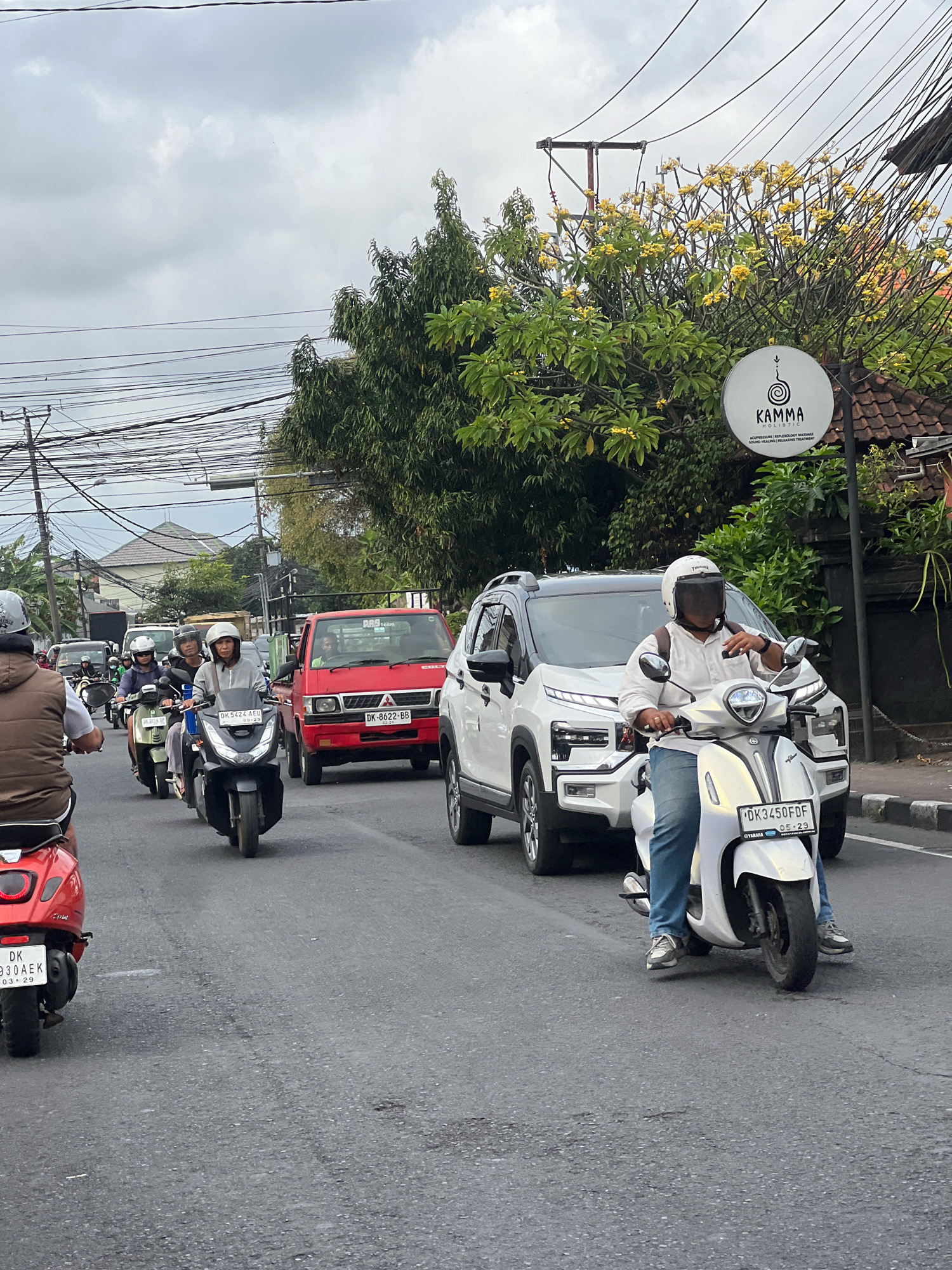 The image depicts a street scene in Denpasar, Indonesia, likely on a moderately overcast day, judging by the cloudy sky. A busy road is filled with vehicles, primarily motorcycles, in a traffic jam. In the foreground, a person wearing a brown vest and helmet rides a red scooter, their legs visible. To their right, a person in a white shirt and blue jeans, wearing a white helmet with a dark visor, rides a white scooter. This scooter has a license plate that reads "DK3450FDF". Behind them, a white SUV with black roof rails is stopped. Further back, a red pickup truck with "DK 8622 BB" license plate is visible, and to its left, a dark-colored scooter with a license plate "DK 5424 AEU" is in motion. Several other motorcycles and their riders are seen further down the road, navigating the traffic. On the right side of the street, there is a building with a tiled roof and a sign for "KAMMA HOLISTIC" which offers services like acupressure and reflexology massage. Trees with yellow flowers are also present, adding a touch of nature to the urban environment. The presence of numerous motorcycles, the style of the license plates (starting with "DK", common in Bali, where Denpasar is located), and the general urban infrastructure suggest the location is Denpasar, Indonesia.