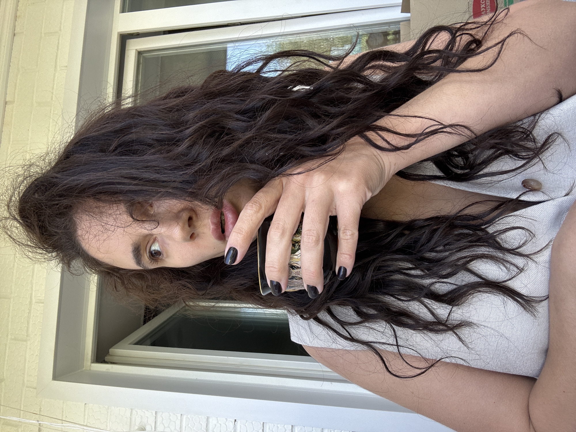The image portrays a woman with long, dark, voluminous, wavy hair, oriented as if looking upwards and to the side with a curious or slightly surprised expression. Her right eye, which is brown, is wide open, and her mouth is slightly ajar, possibly revealing dark lip color. She holds a dark, likely metallic object with intricate gold-colored patterns in her right hand; her fingers are painted with black nail polish. Her dark hair cascades around her head and over her bare arm, resting on a light-colored, possibly white or light grey, textured garment that features a prominent light brown button near her collarbone.
The scene appears to be set indoors or in a semi-enclosed outdoor space, such as a balcony, during daytime, indicated by the natural light. A white-framed window is visible behind her, reflecting outdoor foliage and a bright sky. Beyond the window frame, a light-colored brick wall is discernible. In the background, partially obscured by her hair and arm, a cardboard box shows a fragment of Russian text, "АУКЦ...", suggesting a word like "auction," which aligns with the provided context of Chaikovsky, Russia. The woman's overall demeanor suggests engagement or a moment of candid reaction.