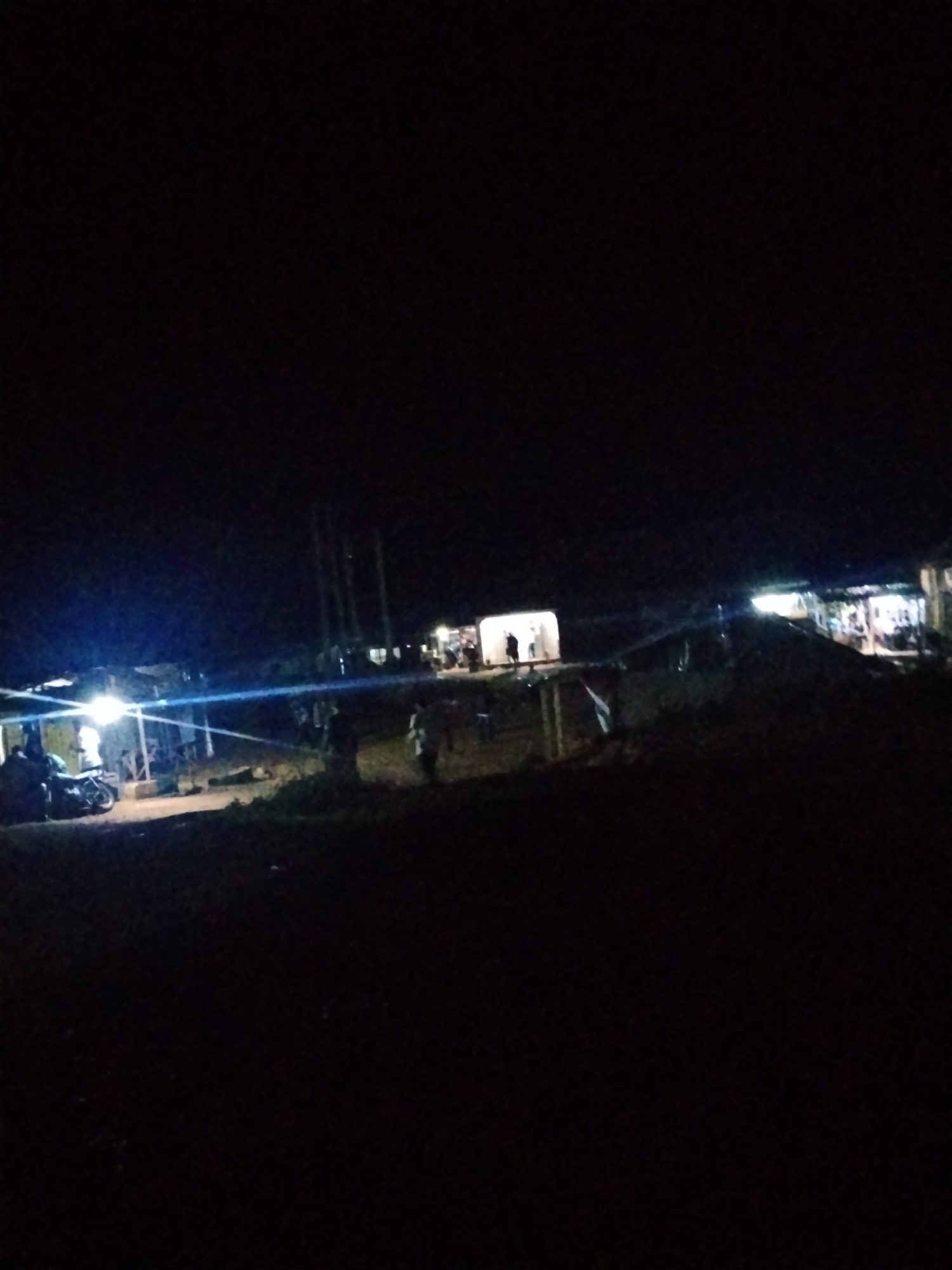 This is a nighttime photograph of an outdoor scene in Durumi, Nigeria. The image shows several illuminated structures, possibly shops or market stalls, with people present. A motorcycle is visible in the foreground to the left. The overall lighting is dim, with bright artificial lights from the structures creating significant glare and lens flare, obscuring details. The environment appears to be a somewhat unpaved area, with some vegetation visible.