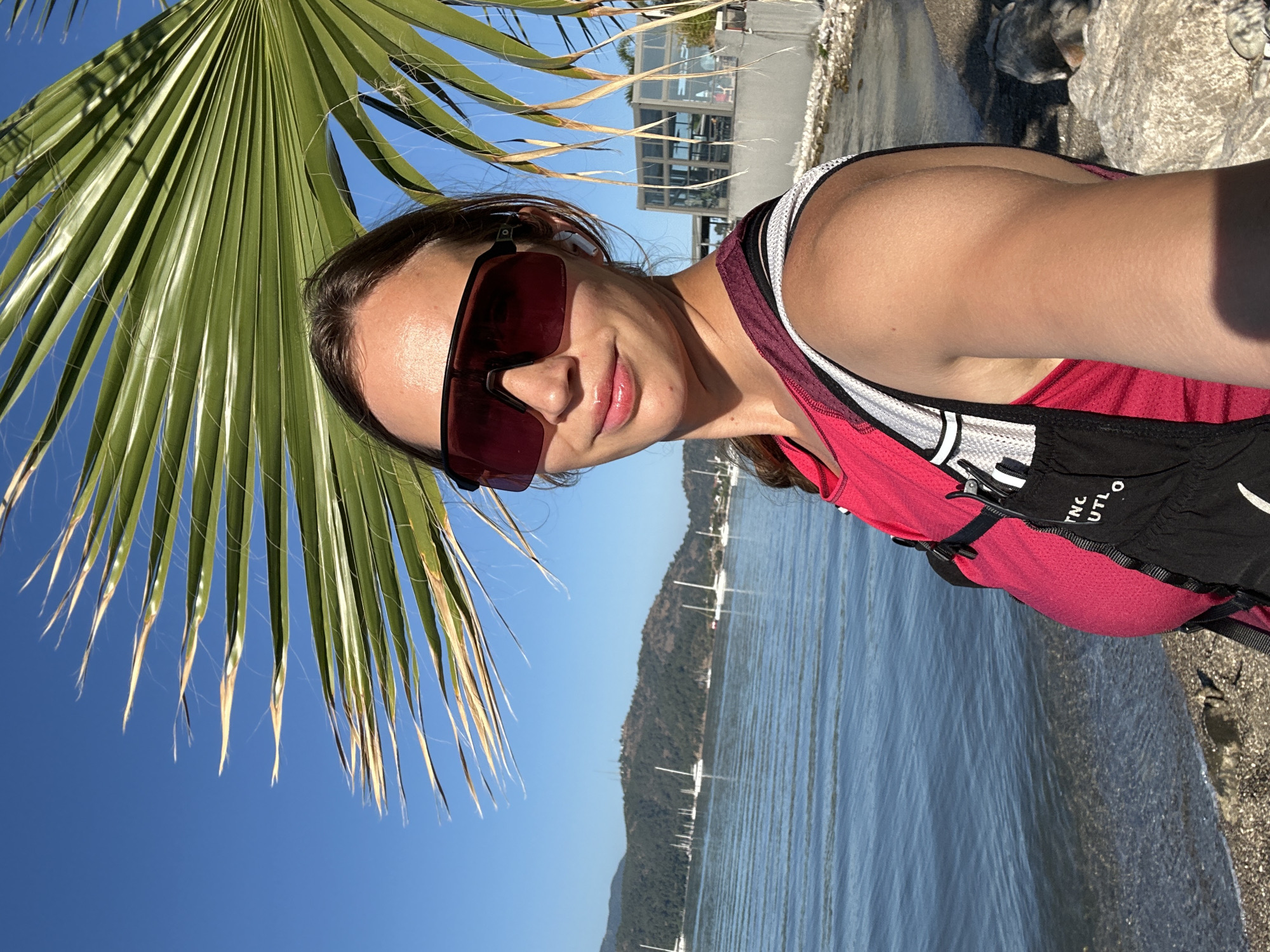 A woman wearing sunglasses, a red tank top, and a black and white hydration pack stands on a beach next to the water. A palm tree is to her left. In the background, there is a body of water with sailboats and a mountainous shoreline. A building is partially visible on the shoreline to the woman's right. The scene is in Fethiye, Turkey.