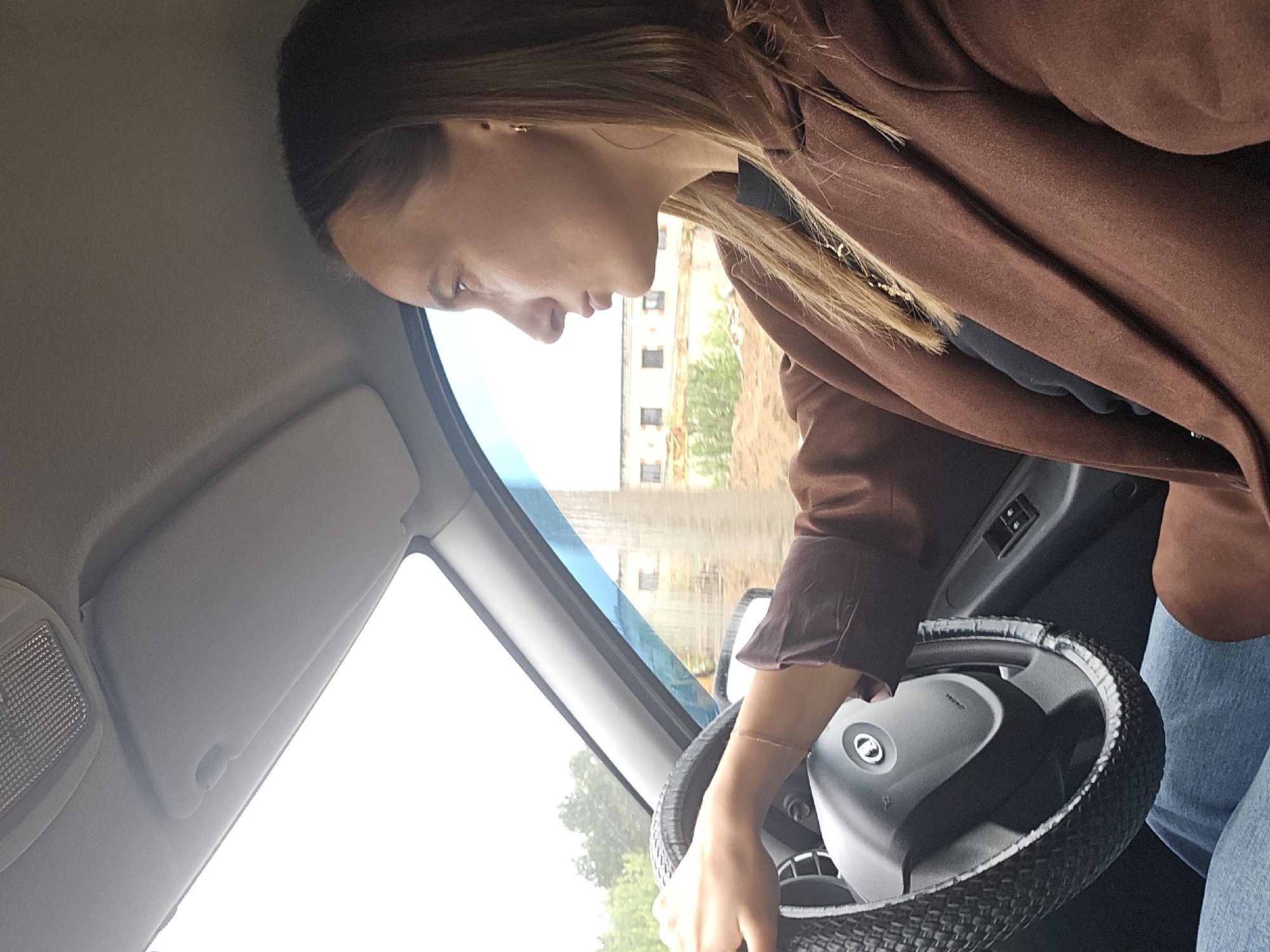 A side-profile shot captures a woman seated in the driver's seat of a car, looking down and slightly forward. She has long, straight brown hair and is wearing a brown jacket over a dark top, along with light blue jeans. A thin gold chain bracelet is visible on her right wrist, which is positioned on the steering wheel. Her hand holds the steering wheel, which is fitted with a dark, textured woven cover. The center of the steering wheel airbag cover shows the word "AIRBAG" and a circular logo.
The car's interior details include the dashboard, a sun visor above, and an overhead light fixture. Through the windshield, a bright, overcast daytime sky is visible, with some distant buildings featuring multiple windows and green trees, indicating an outdoor, possibly urban or suburban, environment. The overall lighting is bright but diffused, characteristic of an overcast day. The woman's expression is neutral and focused. The scene takes place in a vehicle, potentially in Chaikovsky, Russia.