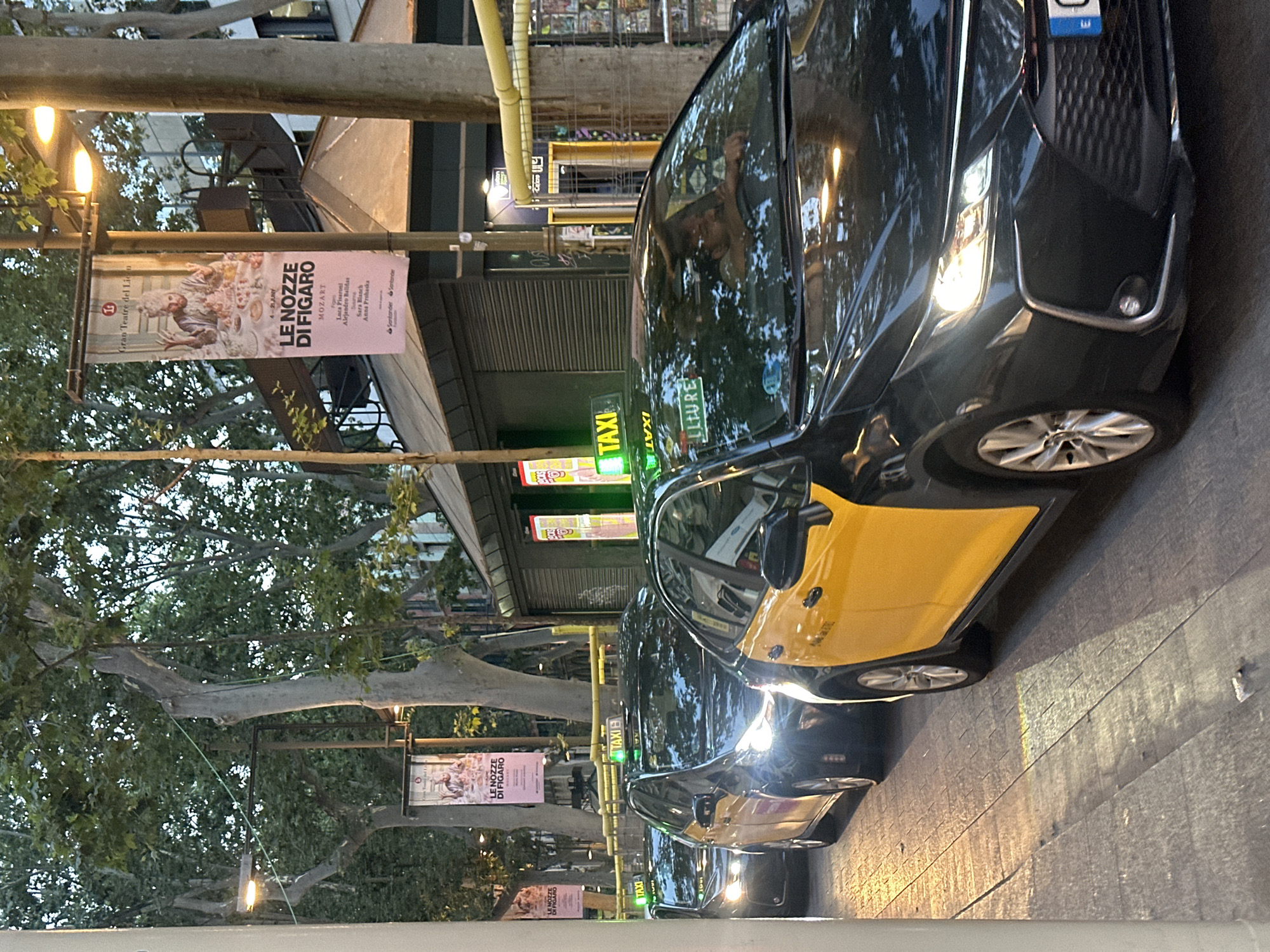 The image depicts a street scene in Barcelona, Spain, likely during the evening or late afternoon, as indicated by the lighting and shadows. Several black and yellow taxis are lined up on what appears to be a paved street. The taxis have illuminated "TAXI" signs on their roofs. One taxi in the foreground is black with a bright yellow rear door and fender. The driver of this taxi is visible through the windshield, looking towards the right. Another taxi behind it is primarily black with a yellow front fender. In the background, there are tall trees with dense foliage and buildings. Banners hanging from lampposts advertise "LE NOZZE DI FIGARO" by MOZART and "Gran Teatre del Liceu". These banners suggest a cultural event or performance is taking place nearby. The environment appears to be urban, with a sidewalk and streetlights visible. The overall atmosphere is that of a busy city street with taxis waiting for passengers.