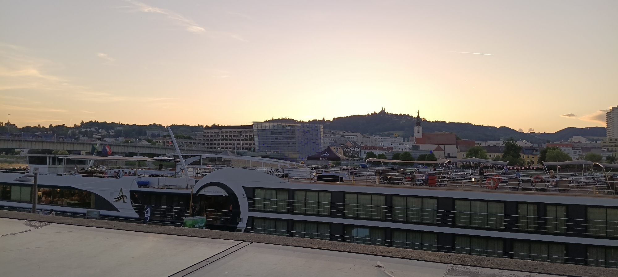 The image captures a scenic sunset over the city of Linz, Austria, with a prominent river cruise ship docked along the Danube River. The sky is a soft gradient of orange and yellow, transitioning to a pale blue at the top, with a faint contrail from an airplane visible.
The cruise ship, identified as the "Avalon Expression" by its visible signage, occupies the foreground and mid-ground. It is a multi-deck vessel with a sleek, modern design. The lower decks feature large, panoramic windows reflecting the dusky sky. The upper decks are equipped with outdoor seating areas, complete with tables and chairs, partially covered by awnings. Several passengers are visible on the upper decks, some seated, engaged in conversation or enjoying the view, while others are standing and moving about. A bicycle is also visible on the upper deck. Two flags, one possibly the Hungarian flag and another green, white, and red flag, are displayed on the upper deck towards the front of the ship.
Behind the cruise ship, the cityscape of Linz unfolds. Buildings of varying architectural styles, including a modern glass-fronted building, a red-roofed church with a steeple, and older residential or office buildings, are nestled against a backdrop of tree-covered hills. A prominent feature on one of the hills is a religious structure with twin spires, characteristic of a church or monastery. A bridge crosses the river in the distance, further accentuating the urban landscape.
The river itself is calm, reflecting the muted colors of the sky. The lighting suggests it is late evening, with the setting sun casting a warm glow over the entire scene. The foreground shows a concrete embankment or walkway, providing a stable vantage point for the photograph.