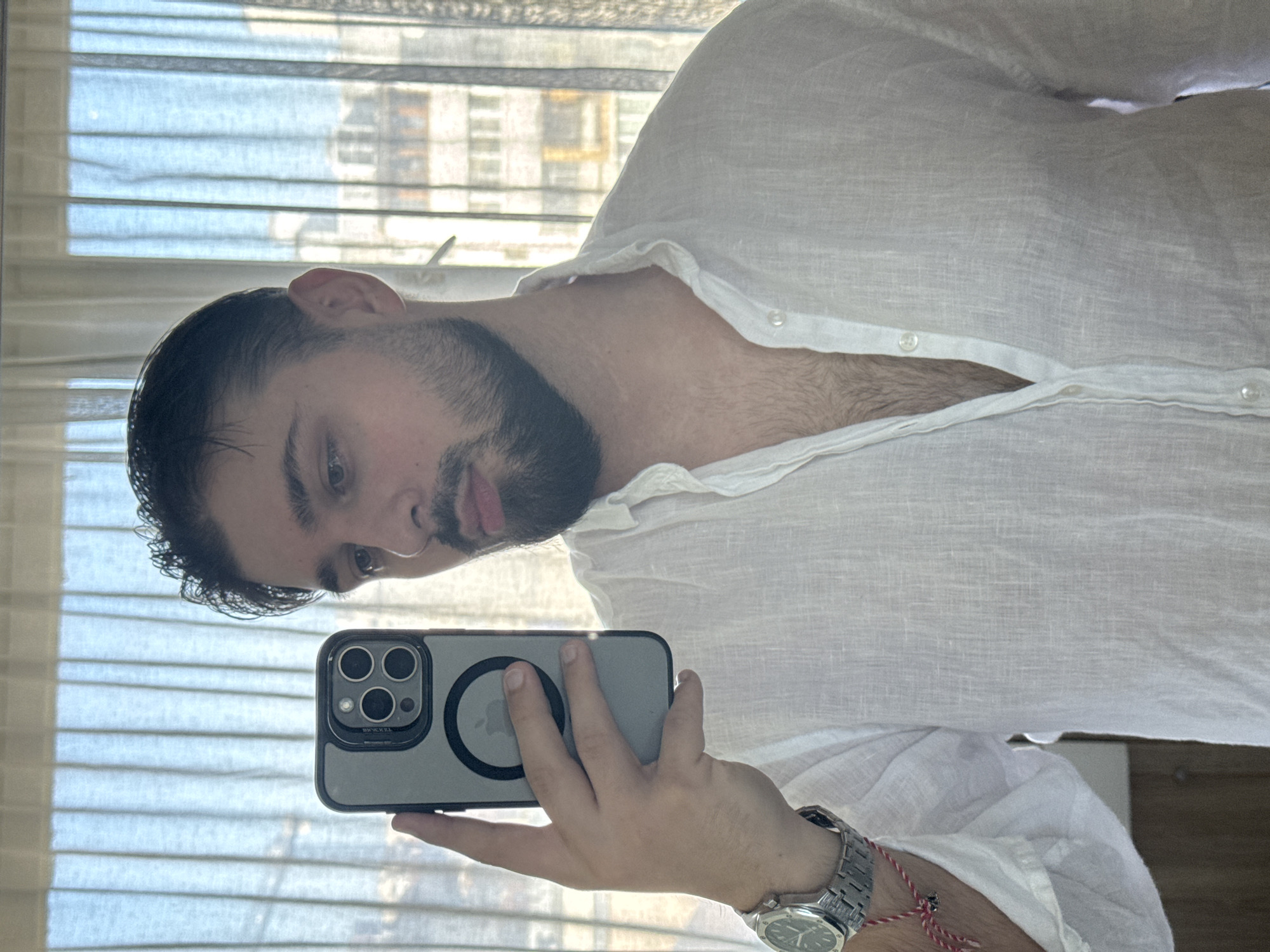 A male individual is depicted in a chest-up selfie, captured indoors in what appears to be Varna, Bulgaria. He has dark hair, styled back, and a neatly trimmed dark beard. He is wearing a white linen shirt, partially unbuttoned to reveal chest hair. His left hand, positioned centrally, holds a grey-cased smartphone (identifiable as an iPhone Pro model with its three prominent camera lenses and a visible MagSafe ring featuring the Apple logo), indicating he is taking the photo. On his left wrist, he wears a silver-toned wristwatch with an octagonal bezel and an integrated bracelet, resembling an Audemars Piguet Royal Oak. Below the watch, a red and white braided string bracelet, a traditional Bulgarian martenitsa, is visible. The background, seen through sheer, light-colored window curtains, reveals an urban landscape with multiple multi-story buildings under a clear sky.