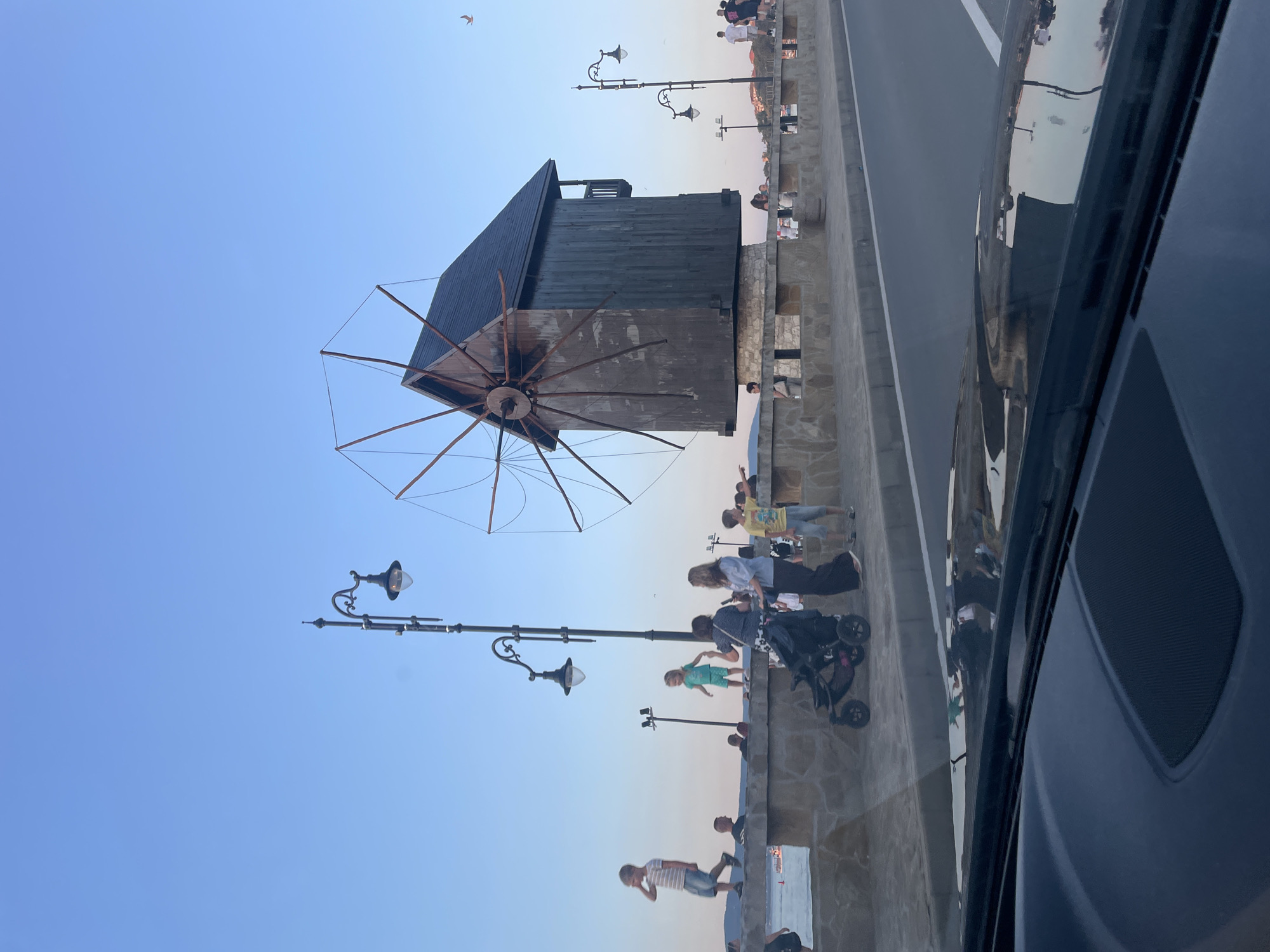 The image depicts a scene in Nesebar, Bulgaria, featuring a wooden windmill situated on a stone embankment overlooking a body of water. Several people are visible walking along the embankment and near the windmill. A child is being pushed in a stroller, and other children are playing or standing on the wall. Street lamps are present along the embankment. The sky is clear and blue, indicating it is likely late afternoon or early evening. The foreground shows the interior of a vehicle, with the dashboard and windshield visible, suggesting the photo was taken from a moving car.