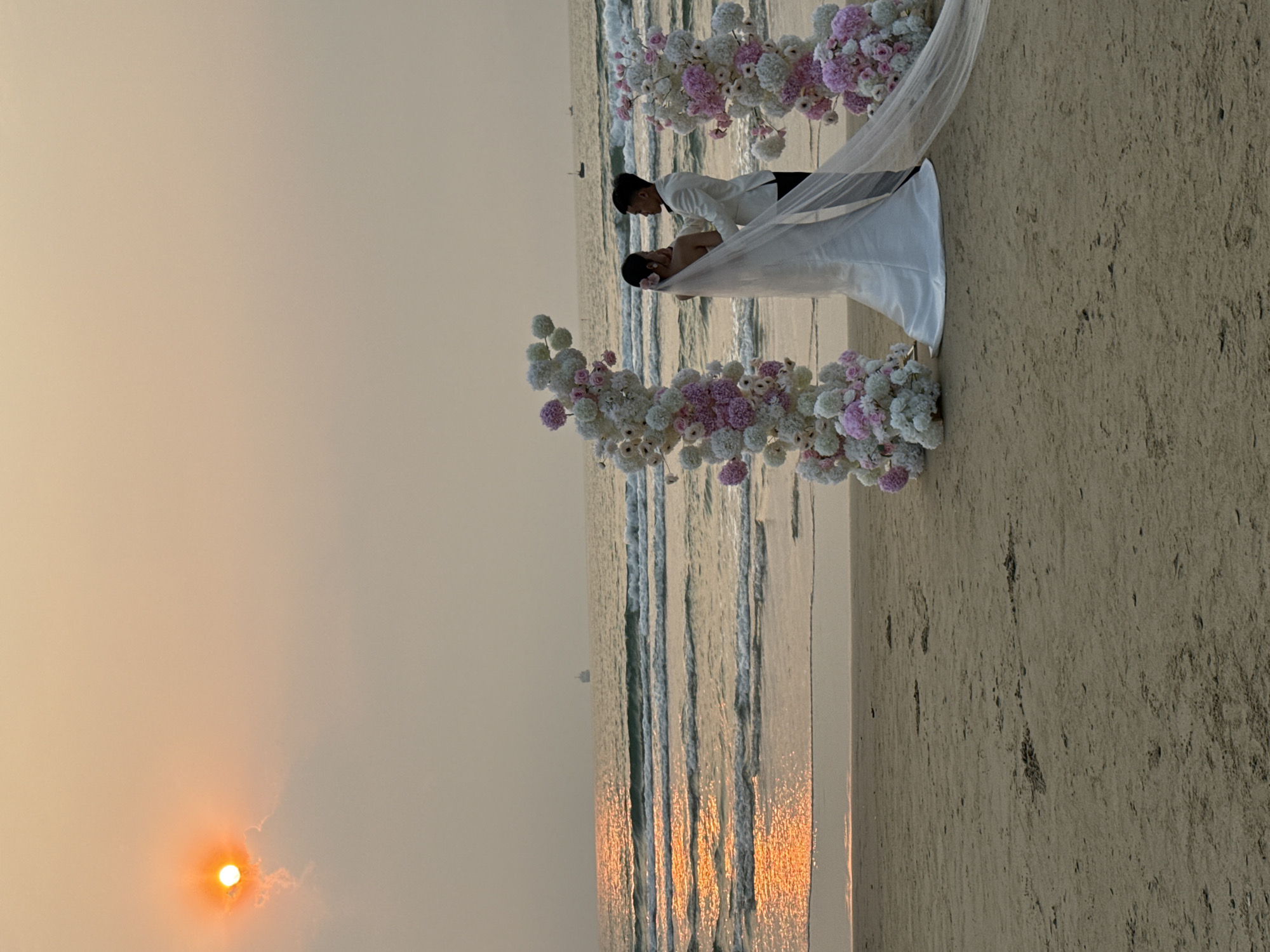 A beach wedding is taking place at sunset on a beach in Đà Nẵng, Vietnam. The couple stands beneath a floral arch of white and purple flowers. The groom is wearing a white jacket, and the bride wears a white dress and veil. The sun is setting over the ocean, casting a warm glow over the scene. The beach is sandy, and the waves are gentle.