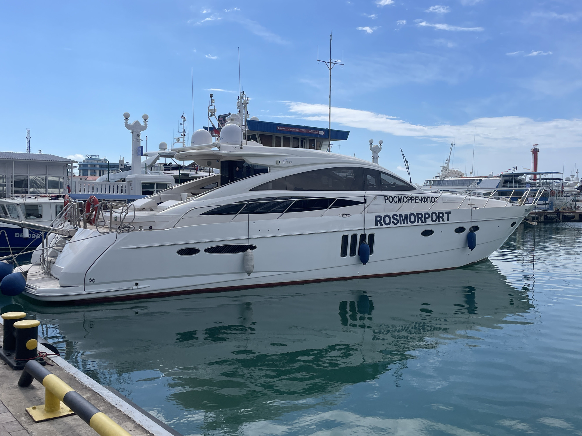A white luxury yacht with the word "ROSMORPORT" written in black letters on its hull is docked in a harbor. The yacht has a sleek design with tinted windows and several antennas on its roof. In the background, other boats and harbor structures are visible under a bright blue sky with scattered white clouds. The water is calm and reflects the yacht and the sky. The location is the Sochi Sea Port in Sochi, Russia. There are no people visible in the image.