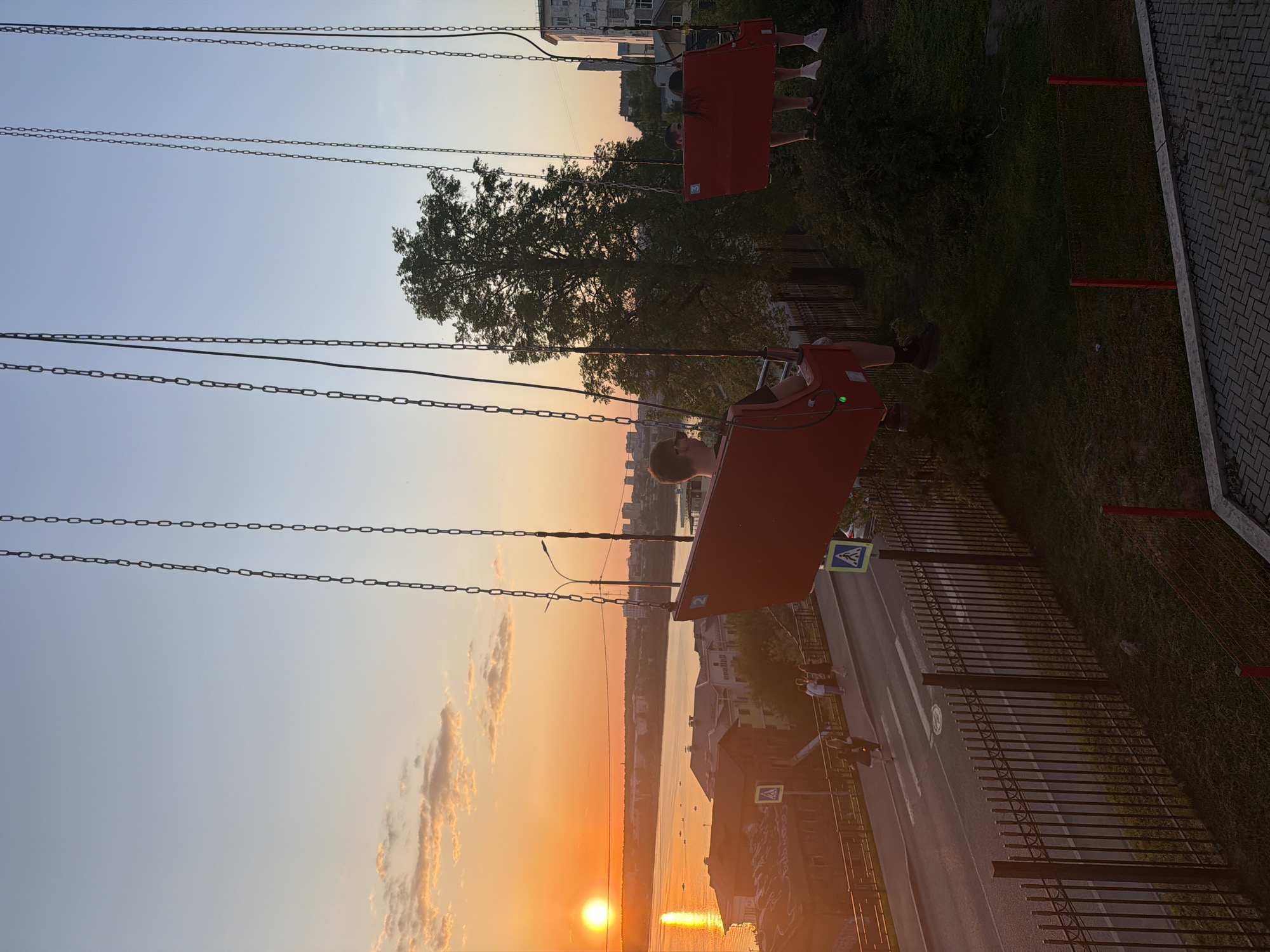 The image captures a scene from a swing ride overlooking a cityscape, likely in Izhevsk, Russia, based on the architectural style and urban layout. The swing ride, consisting of seats suspended by chains, features occupants in motion. In the background, a city with buildings and a body of water reflects the setting sun. A street with a pedestrian crossing and fencing runs parallel to the river. The overall environment suggests a park or recreational area near a developed urban zone during a sunset.