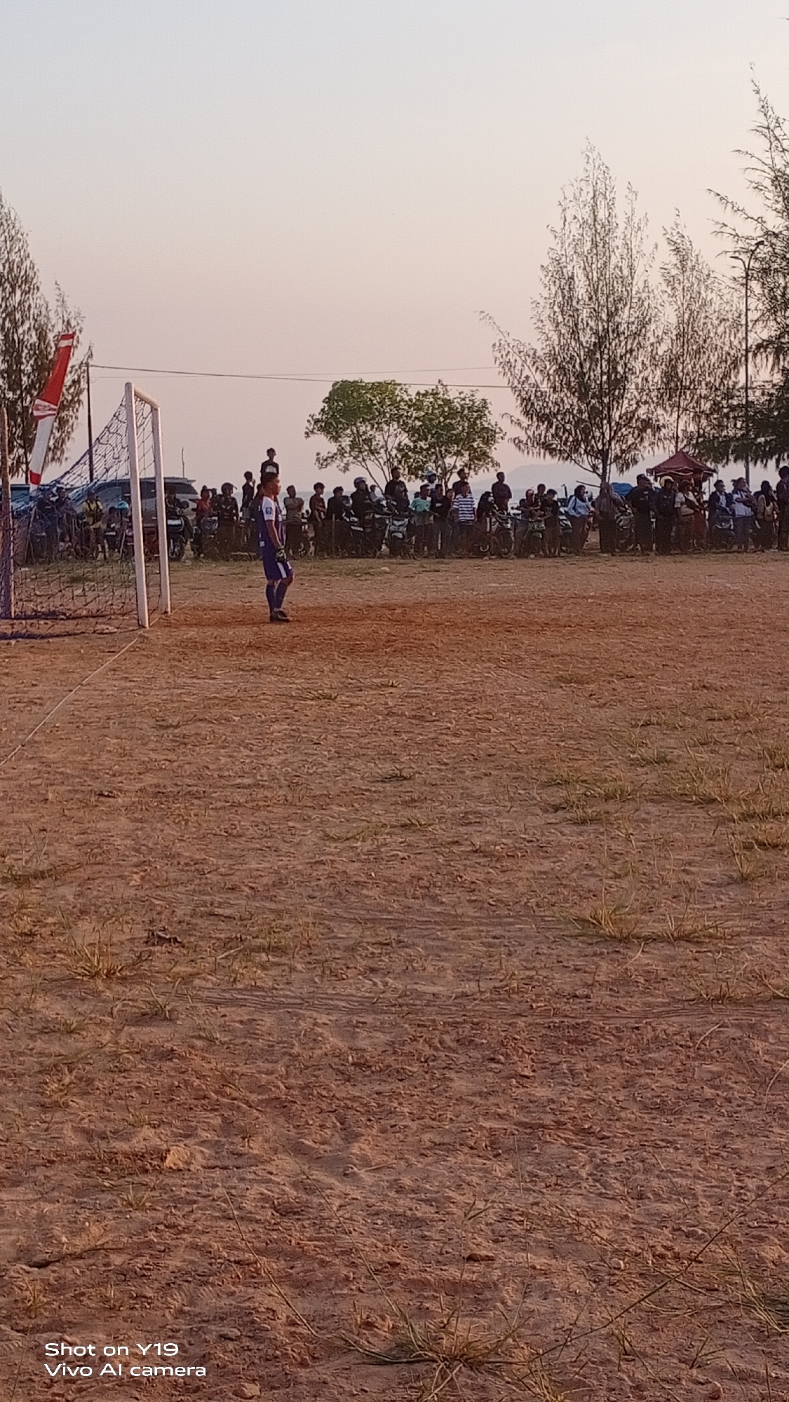 The image captures a soccer game in progress on a dirt field, likely located near the coast in Bajo, Indonesia. A lone player in a blue and white uniform stands on the field, facing away from the camera, near a soccer goal with a white net. In the background, a crowd of spectators stands behind a line of parked motorcycles and a few cars. Trees, including some tall, slender ones and a larger, bushier one, are visible behind the crowd, with a pale sky overhead. The ground is primarily composed of reddish-brown dirt with patches of dry grass. The overall scene suggests a local community event taking place outdoors.