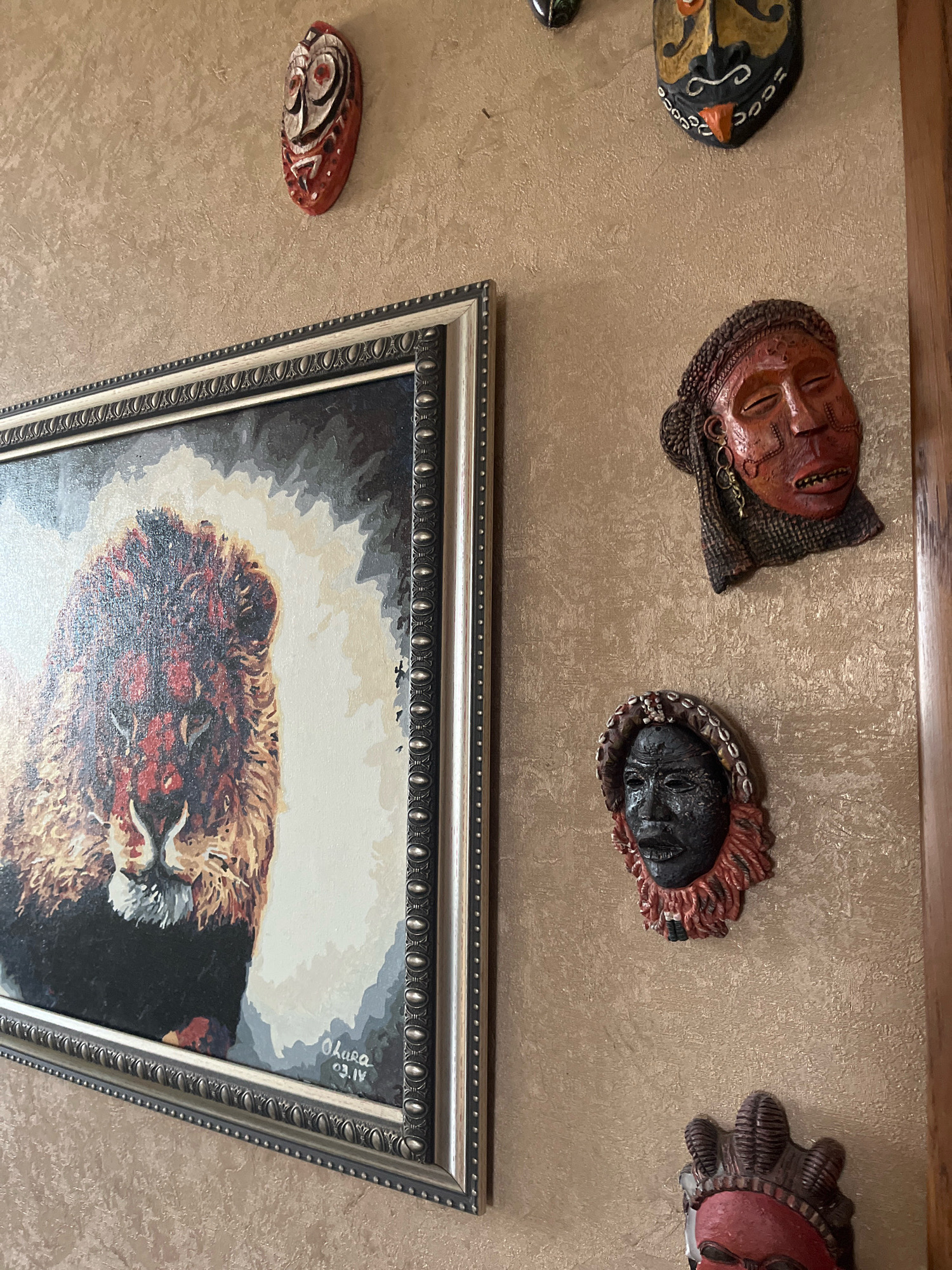 The image shows a wall decorated with several African masks and a framed painting of a lion. The wall has a textured, beige finish. The painting is in a silver frame with intricate detailing. The masks are of various sizes and designs, with some featuring intricate carvings and bright colors. One mask is red and white with circular eye details. Another is black with a red fringe and bead detailing around the forehead. A third mask is red and brown with textured hair elements. A fourth mask is red and black with a distinctive headdress. A fifth mask, at the top right, is black with yellow and orange accents and a prominent orange nose and tongue. The sixth mask, partially visible at the bottom right, is red with a dark headdress. The painting appears to be a portrait of a lion's face, rendered in a bold, impressionistic style with prominent reds and browns in its mane and face. The signature "Ohara 03.IV" is visible in the lower right corner of the painting. The overall impression is of a decorated interior wall, likely in a home or gallery, displaying cultural artifacts and artwork. There is no information in the image to suggest a location in Vladimirovka, Russia.
