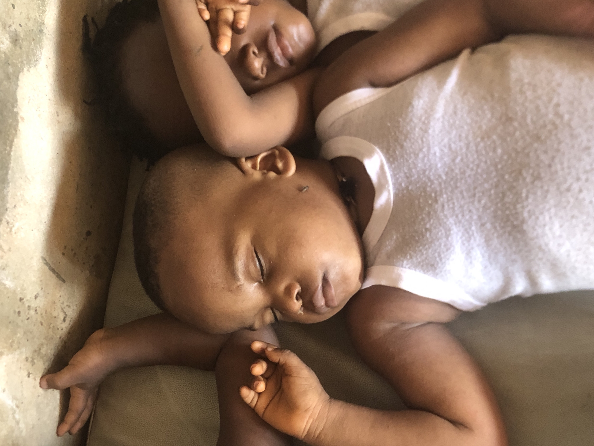 Two young children are sleeping side-by-side, their faces close together on a textured surface, possibly a couch. The child in the foreground is on their side, eyes closed, with one arm resting outward and a hand partially open. The child in the background is also on their side, with their face partially obscured by their arm and hand covering their eyes. Both children are wearing white tank tops. The environment appears to be indoors, with a rough, textured wall visible in the background. The scene is lit from above, casting shadows. The image was captured in Ukwu-Abwa, Nigeria.