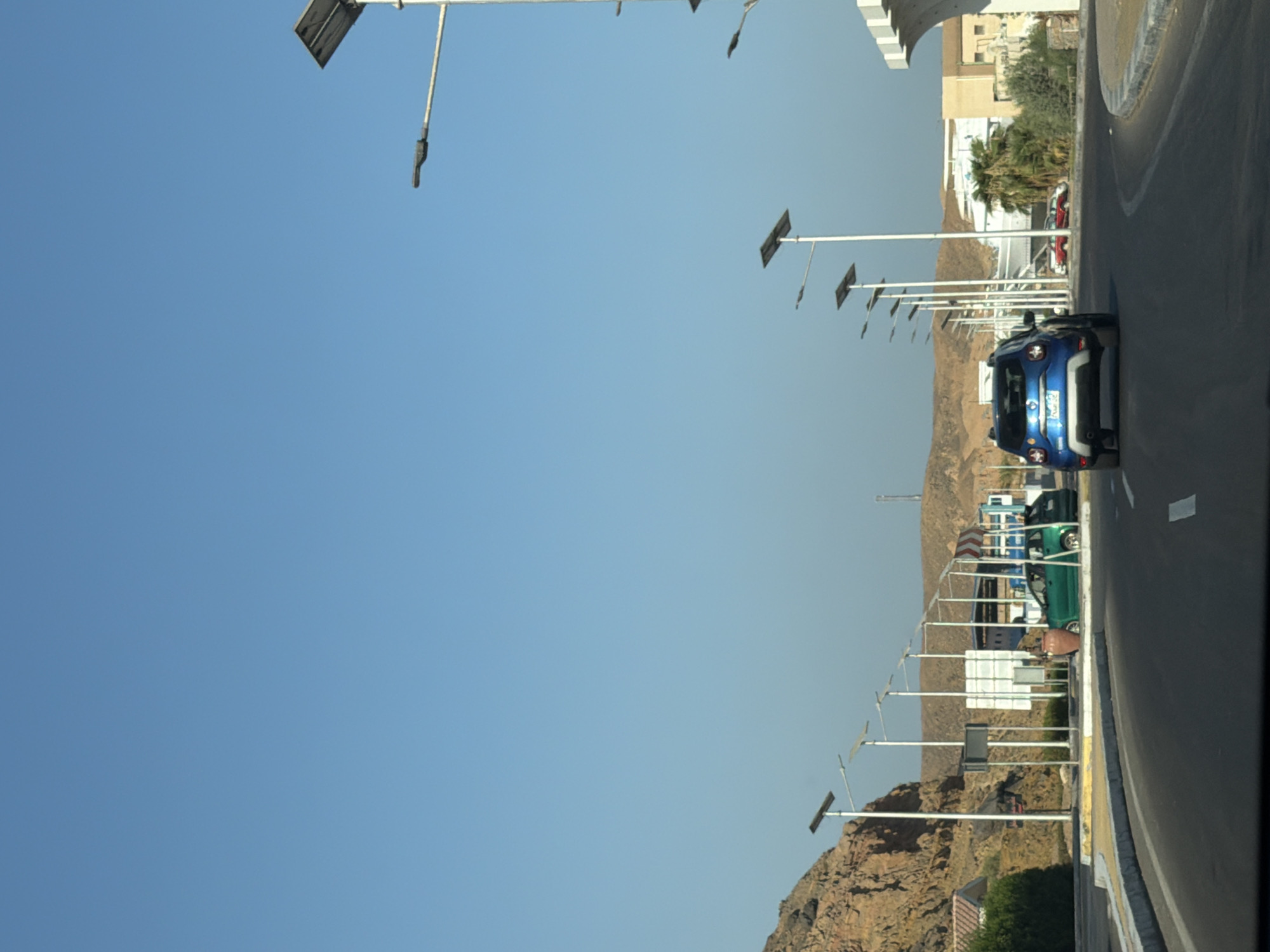 The image depicts a street scene in Sharm El Sheikh, Egypt, under a clear blue sky. In the foreground, a road curves to the right, with a blue SUV driving away from the viewer. The road is bordered by a grey curb and a yellow line. To the left of the road, a rocky, barren hillside rises. Along the side of the road, a series of streetlights with solar panels are visible, extending into the distance.
Further down the road, a green car is parked. Behind the green car, there are buildings and palm trees. There are also some signs and what appears to be a bus shelter or a covered waiting area. The overall environment suggests a desert landscape with urban infrastructure. No people are clearly visible in the image.