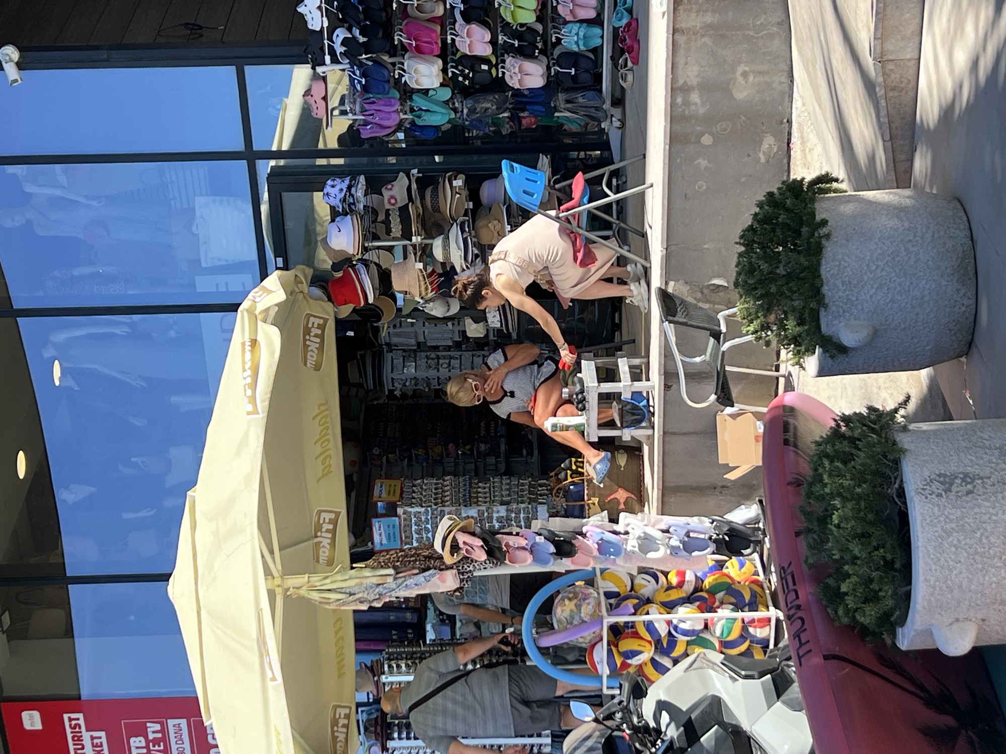 The image depicts a street-level view of a shop in Rafailovići, Montenegro, specializing in beachwear and accessories. The shop features a large glass storefront displaying mannequins, and a prominent yellow awning shades an outdoor display. Two women are present: one seated on a low stool, wearing sunglasses and a patterned top, is handling a drink bottle. The other woman, seated on a blue plastic chair, wears a light-colored dress and is reaching towards a red object. A variety of items are displayed, including hats stacked on shelves, colorful flip-flops and sandals arranged on racks, and inflatable beach balls. In the foreground, a red paddleboard with "THUNDER" branding is visible, along with two potted plants. The overall scene suggests a commercial area catering to tourists in a sunny, outdoor environment.