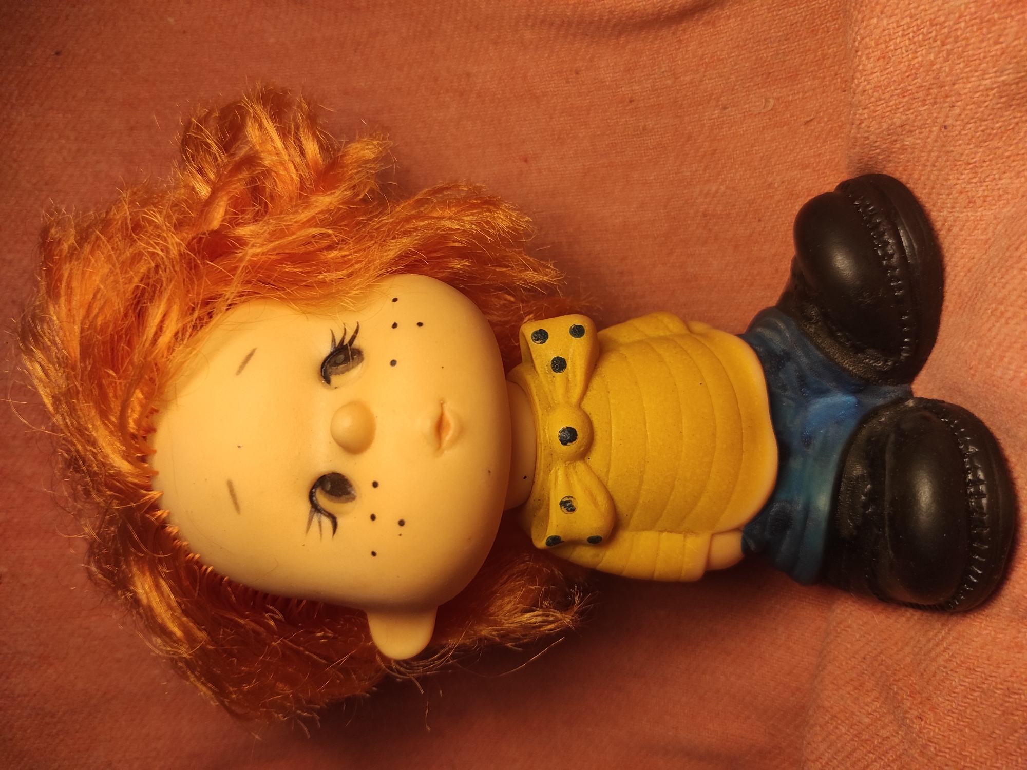 The image shows a doll with bright orange, curly hair, a yellow shirt with a blue polka dot bow tie, and blue pants. The doll has a round face with painted eyelashes and freckles, and a small nose and mouth. It is sitting on a textured, pinkish-orange surface. There is no indication of the doll's location in Chernihiv, Ukraine.