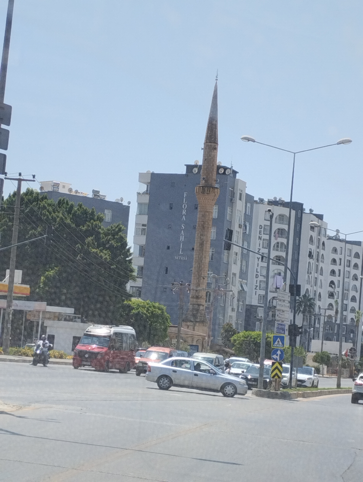The image captures a street scene in Mezitli, Turkey, on a clear, sunny day. A prominent minaret, characteristic of Islamic architecture, stands tall against the bright sky, with a pointed spire. Beside it, a modern, multi-story building displays the text "FLORA SAHİL SITESİ" vertically on its facade. Another adjacent building, also multi-story, has "DENİZ YILDIZI" written vertically.
In the foreground, a traffic intersection is visible. Several vehicles are present, including a red and white minibus, a silver sedan, and other cars. A person is seen riding a motorcycle. Traffic lights and street signs, including directional arrows, are also visible at the intersection. Lush green trees line the street, interspersed with the urban structures. The overall environment suggests a typical day in a bustling Turkish city.