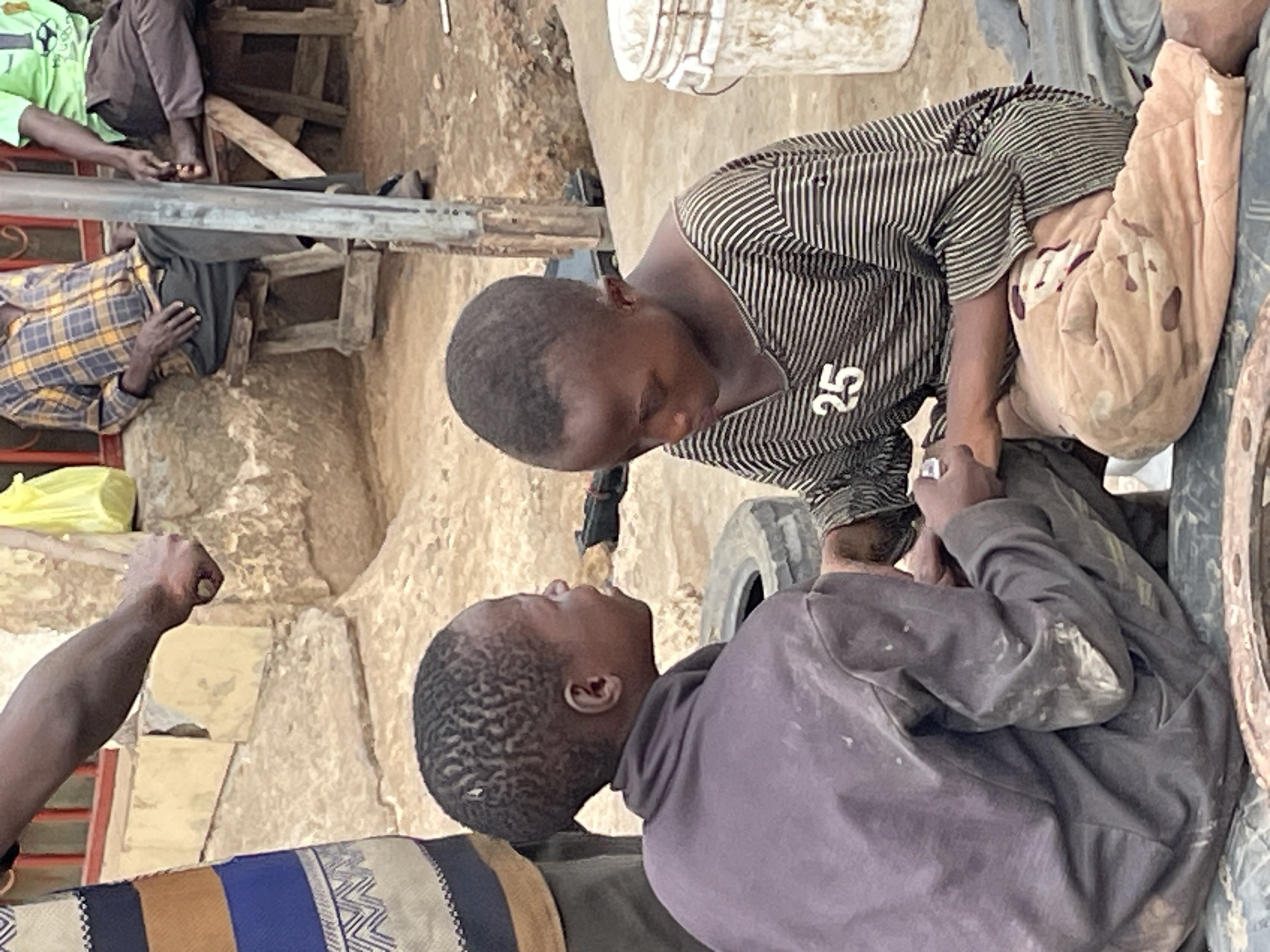 This photograph, presented on its side, captures a focused interaction between two young individuals in an outdoor, informal setting in Jos, Nigeria. In the foreground, two boys are seen face-to-face, their hands clasped or touching centrally.
The boy positioned lower in the frame has short, curly dark hair and wears a dark grey or purple hoodie, which appears smudged with white marks, possibly from dust or paint. He looks up intently at the other boy. This second boy, positioned above him, has very short dark hair and is dressed in a striped shirt, featuring black and white or dark and light grey stripes, with the number "25" visibly printed on his sleeve. He looks down, meeting the gaze of the first boy. Their direct eye contact and clasped hands suggest a moment of deep connection, a handshake, or a shared gesture of focus or comfort.
The environment is rustic and appears to be a dry, dusty area with light brown ground. Various makeshift elements define the scene. In the background, other individuals are partially visible, seated on rudimentary wooden structures. One person wears a light green shirt, while another is in a yellow and dark plaid shirt. Objects such as a white bucket, a metal pole, and tires (which seem to be used as parts of the structure or seating) are also present. The overall atmosphere suggests a casual community gathering or a break from activities in a utilitarian outdoor space under natural daylight conditions.