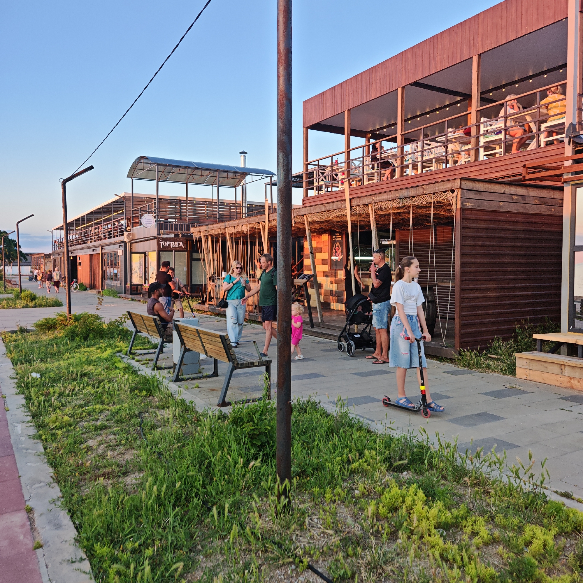 The image captures an outdoor scene with a clear sky and a row of buildings that appear to be restaurants or shops, possibly along a boardwalk or waterfront. The architecture of the buildings is modern, featuring wooden and metal structures with open-air seating areas on multiple levels.
In the foreground, a paved walkway is lined with benches and patches of green grass and weeds. A young girl with dark hair, wearing a white t-shirt and a denim skirt, is riding a scooter. She is facing to the left and appears to be focused on her ride.
Further back, several adults are visible. A woman in a teal top and light-colored pants is standing and holding a bottle. A man in a green shirt and dark shorts is walking next to her, and a young child in a pink outfit is near them. Another man in a black t-shirt and denim shorts is standing and smoking, with a stroller beside him. People are seated at tables on the upper level of one of the buildings.
The setting suggests a public space, possibly a tourist or recreational area, during the late afternoon or early evening, given the warm lighting. The overall atmosphere appears relaxed and casual.