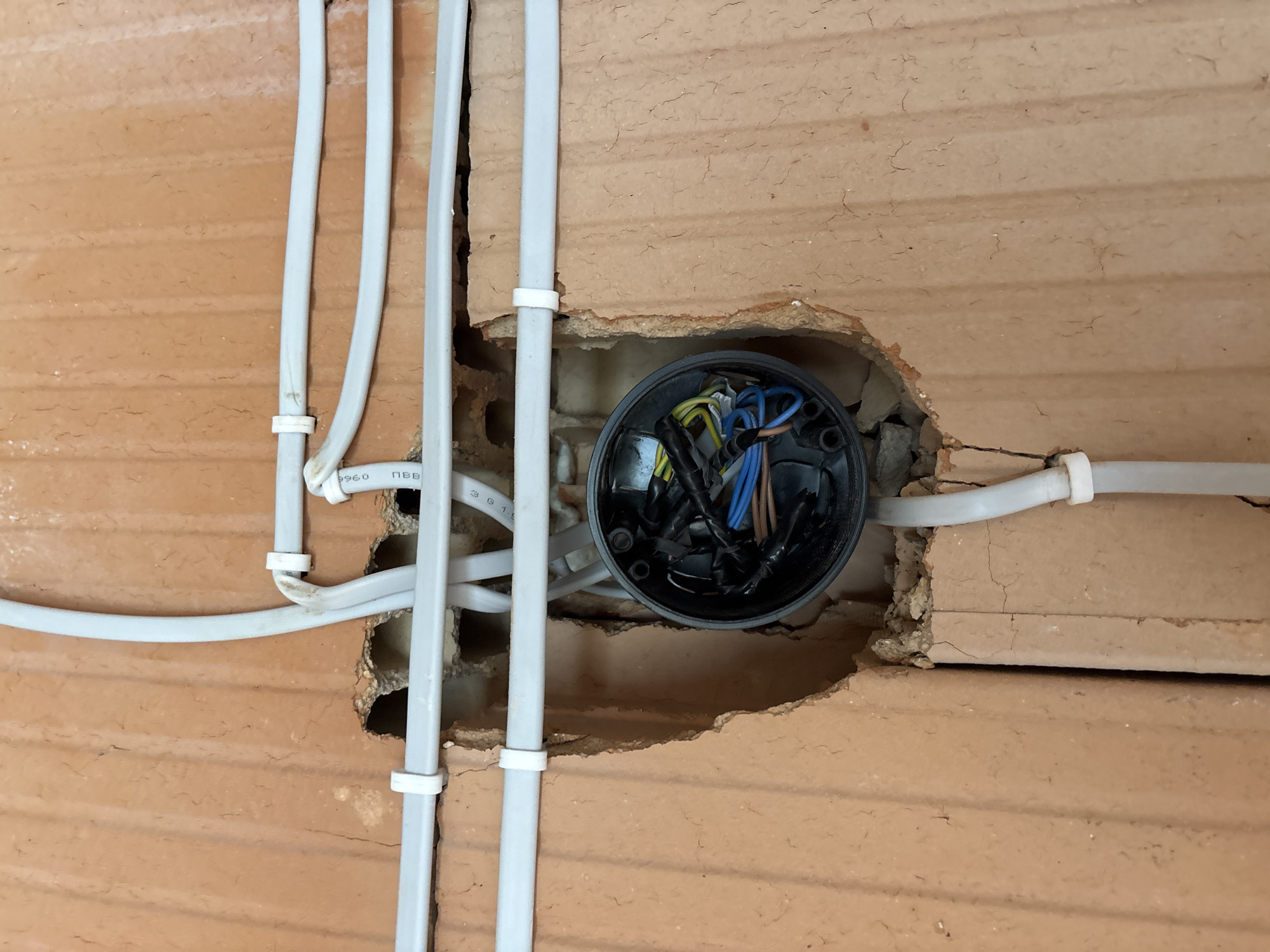 The image shows a partially constructed electrical installation in a brick wall. A black electrical junction box is recessed within a roughly circular hole in the wall. Several grey electrical cables, secured to the wall with white plastic fasteners, feed into the junction box. The wires inside the junction box are visible and are color-coded. The wall appears to be made of red clay bricks. The image was taken in Burgas, Bulgaria.