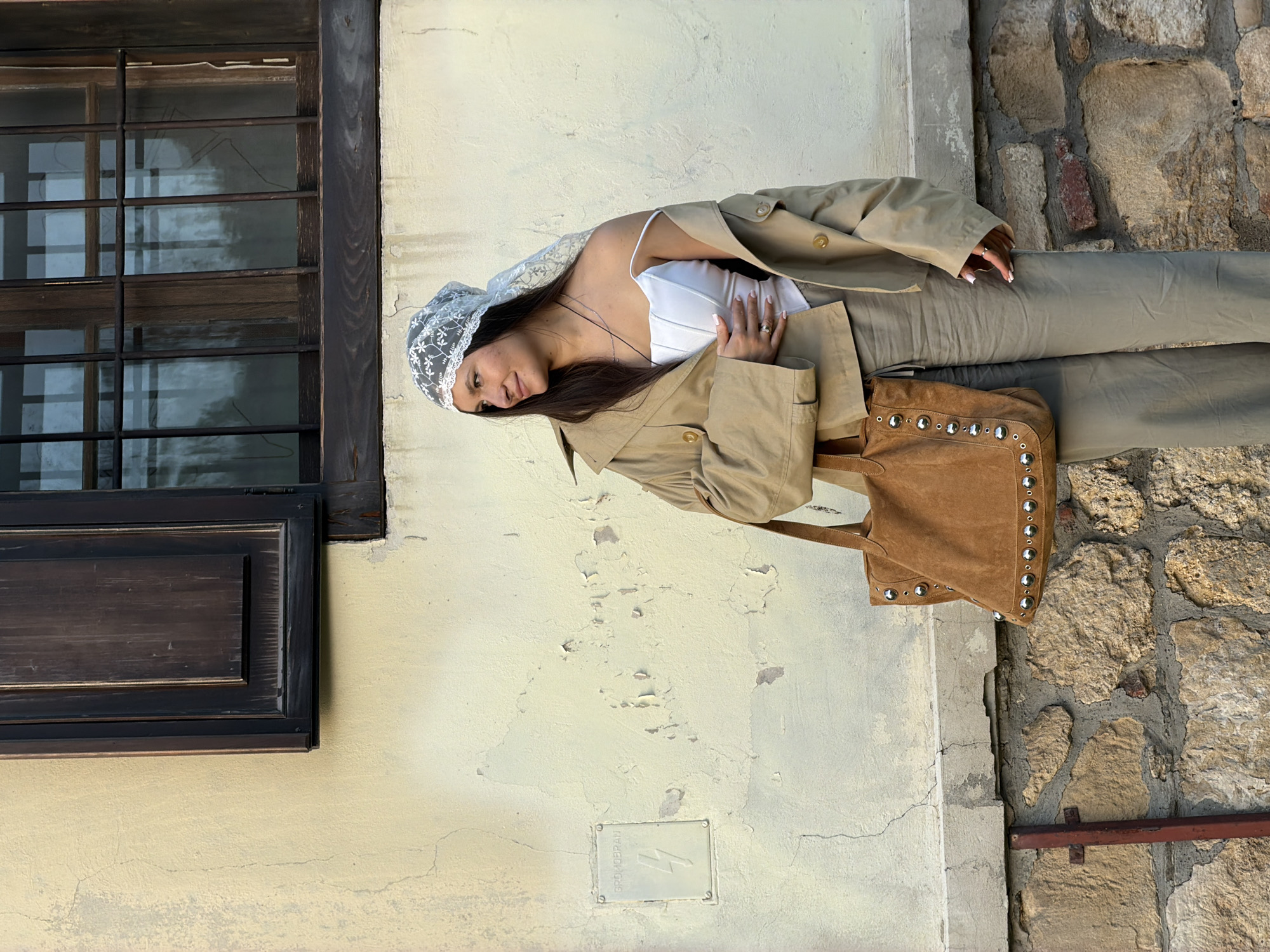 A female figure is angled left in a full-body shot against a backdrop of varied architectural surfaces. The scene includes a window with dark wooden frames, a beige wall with an electrical panel, and a stone wall. The woman is wearing a white top, a light trench coat, beige pants, and a lace head covering. She carries a studded suede tote bag. The setting appears to be in Belgrade, Serbia.