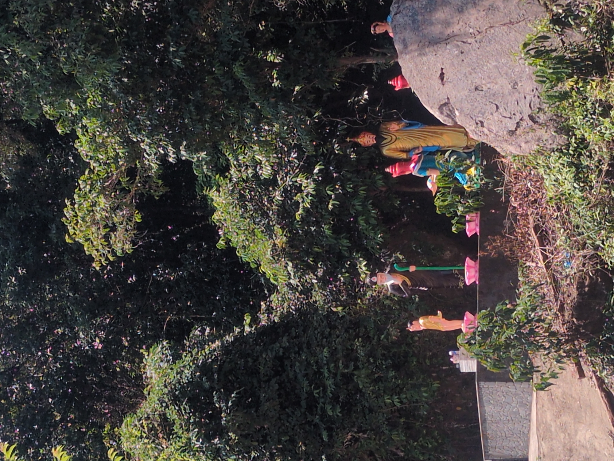 The image shows a statue installation in Phú Quốc, Vietnam. The installation consists of several brightly colored religious figures, including statues of people in traditional attire, arranged along a rock formation or hillside amidst dense green vegetation. Pink candle holders are placed at various points along the path where the statues are located. The setting appears to be a garden or a wooded area near a stone wall or walkway.