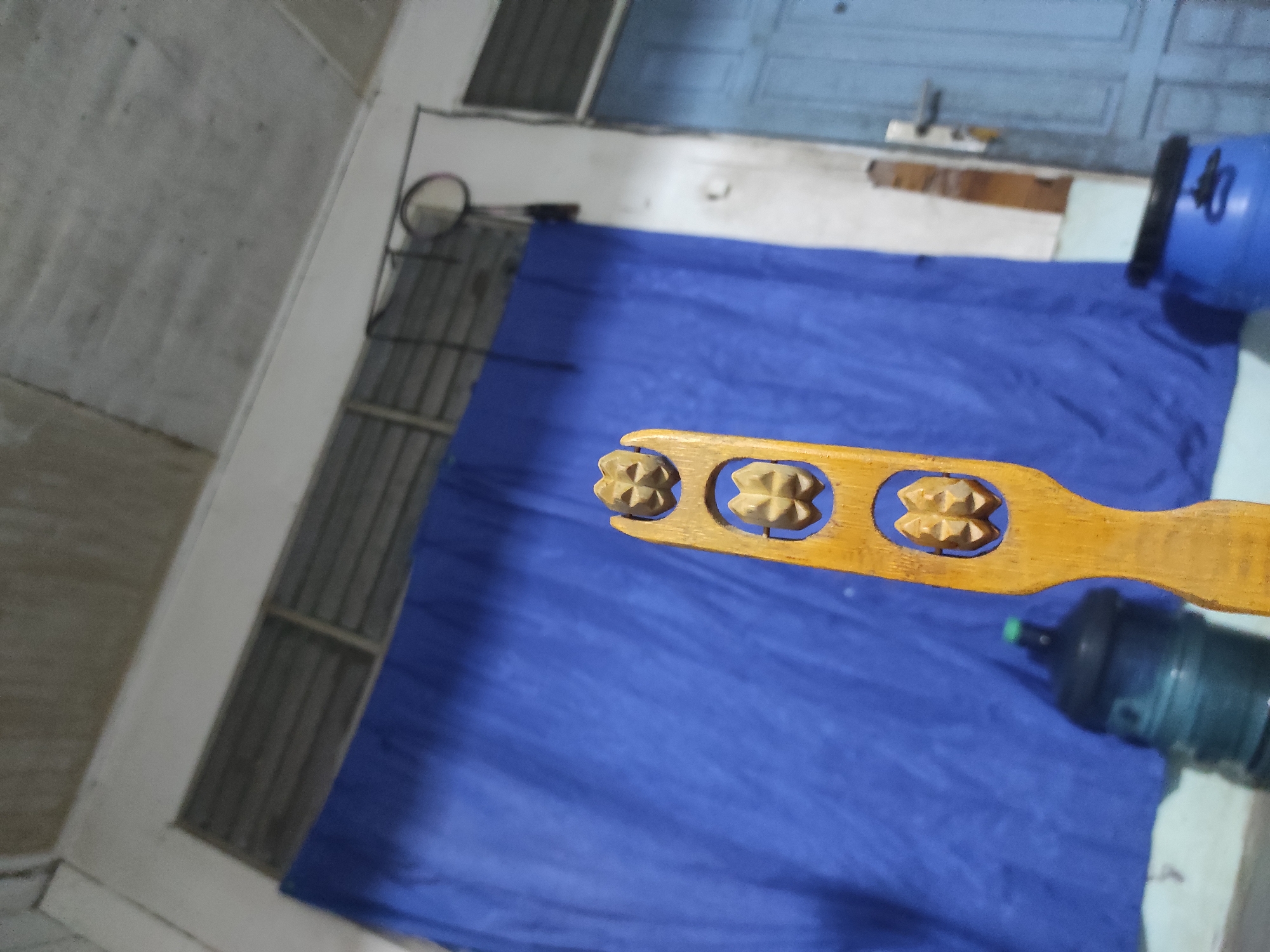 A wooden massage roller with a natural finish is the central subject of the image, positioned horizontally across the frame. It features three sections, each with two spiked, star-shaped rollers designed for massage.
In the background, a large blue tarp or piece of fabric covers most of the lower and central area. On the left, a grey concrete wall is visible, along with what appears to be a window with metal bars. Above the tarp, a white wall and a blue door are discernable. A badminton racket frame is also visible near the white wall. To the right of the tarp, two large water bottles are present: one blue, partially cropped, and another greenish-black bottle further down the frame.
The scene appears to be an indoor or covered area, possibly a utility space or a casual room. There is no human activity or visible text. The lighting suggests an indoor environment. The setting indicates a practical, functional space in what is identified as Uteungeulinggang, Indonesia.