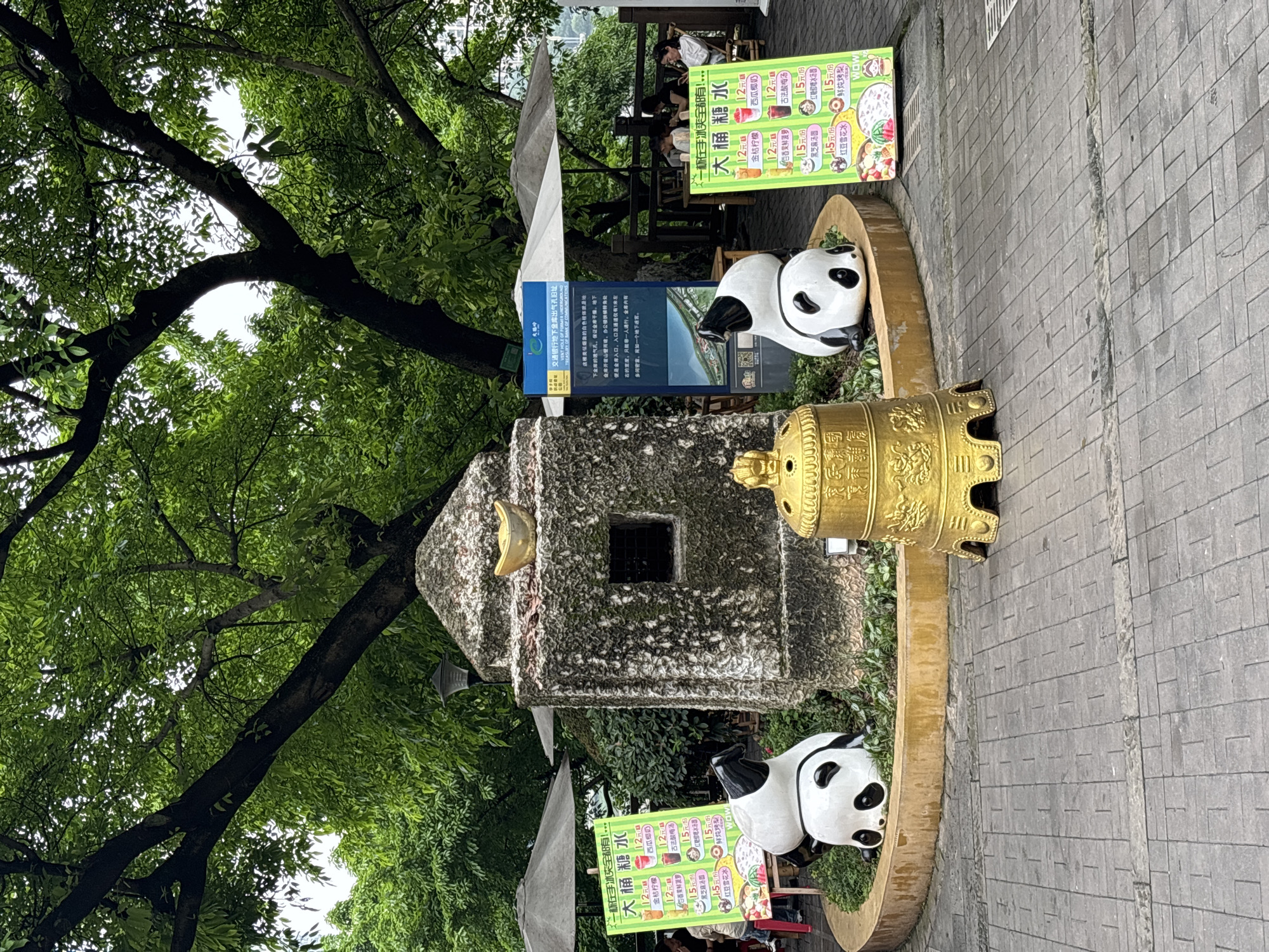 The image captures a scene on a paved walkway in Yuzhong District, Chongqing, China, marked by a combination of cultural and commercial elements. Dominating the view are two panda statues positioned on either side of a stone monument, possibly an old ventilation shaft for the "Traffic Bank underground treasury," as indicated by a sign with Chinese writing. A golden bell and two menu signs advertising "Big Bucket Sugar Water" stand nearby. The background includes a tree that partially covers the top left portion of the image. In the background, there are some patrons sitting on a bench at a small table. The photograph is shot at a slightly vertical angle. The foreground is filled with gray tiles.