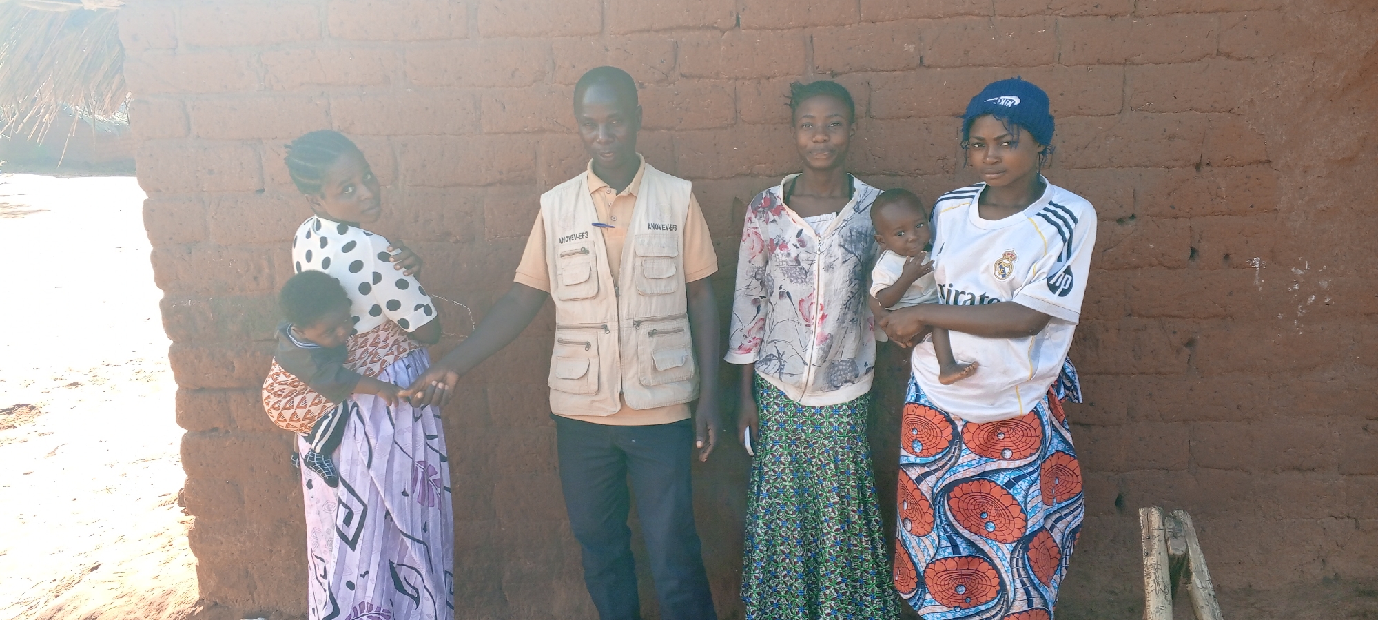 In Ngalula, Democratic Republic of the Congo, four adults and two children stand in front of a mud brick building. A man in a tan vest labeled "ANOVEV-EF3" holds hands with a woman in a polka dot shirt, who carries a child in a sling. Another woman wearing a floral blouse stands next to them. To the right, a woman in a Real Madrid jersey and a blue knit cap holds a small child. The building behind them appears to be a residence.