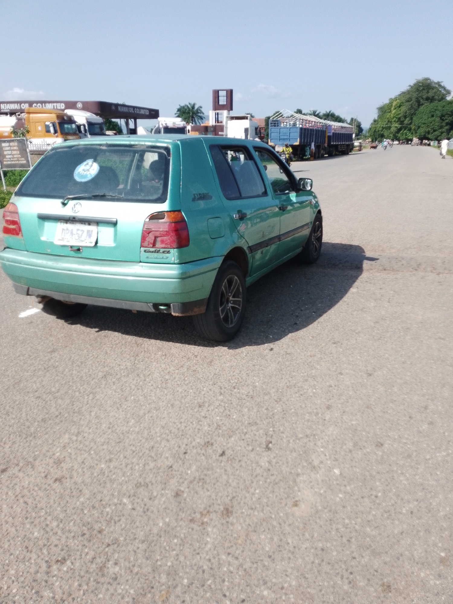This image captures a serene outdoor scene in Serti, Nigeria, likely during the daytime given the bright sunlight casting distinct shadows. The foreground is dominated by a faded green Volkswagen Golf hatchback, parked on a paved road. Its rear bumper shows signs of wear, and a license plate with the text "DPA-57 N" is clearly visible.
In the background, a prominent "NJAWAI OIL CO LIMITED" sign arches over a gas station, hinting at a commercial area. Several large trucks are parked or moving along the road, some carrying goods and appearing to be part of a busy transport route. A few people are visible on motorcycles and on foot, adding a sense of everyday life to the scene. Lush green trees line the right side of the road, suggesting a tropical or sub-tropical environment. The sky is a clear, bright blue with a few scattered clouds. The overall impression is one of a typical day on a highway in a Nigerian town, with commercial activity and local transport in full swing. The text visible includes "NJAWAI OIL CO LIMITED", "WORSHIP WITH US", "LIGHT (ASK)", "BAPTIST CHURCH", "SERTIA", and the license plate information.