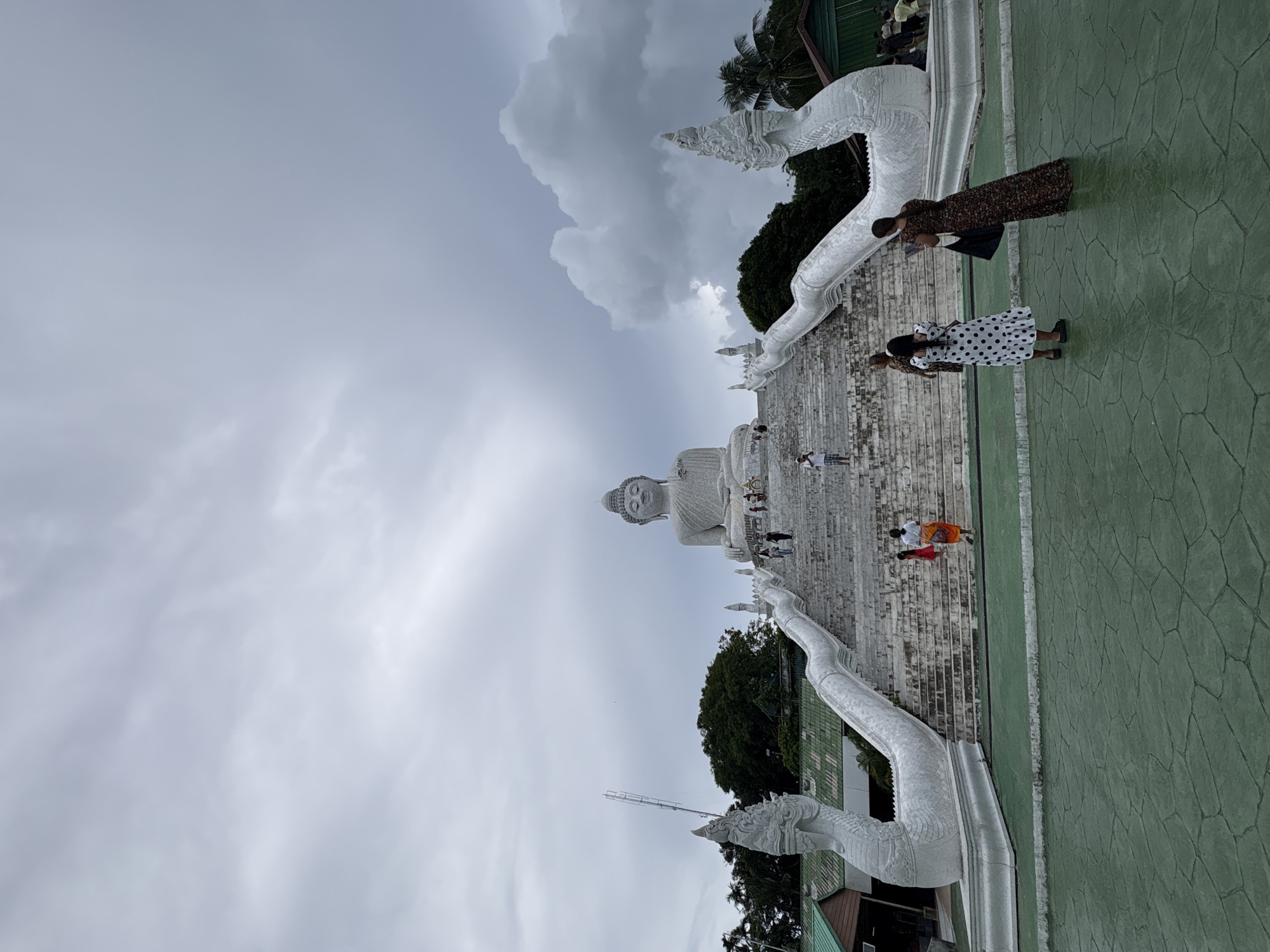 The image depicts the Big Buddha statue in Rawai, Phuket, Thailand. A large, white marble Buddha statue sits atop a hill, overlooking a lush green landscape. The statue is approached by a long, wide staircase, flanked by ornate white serpent sculptures that curl upwards. Several tourists are seen ascending the stairs, some taking photographs. The sky above is overcast with dramatic, dark clouds. The ground in the foreground is a textured green surface.